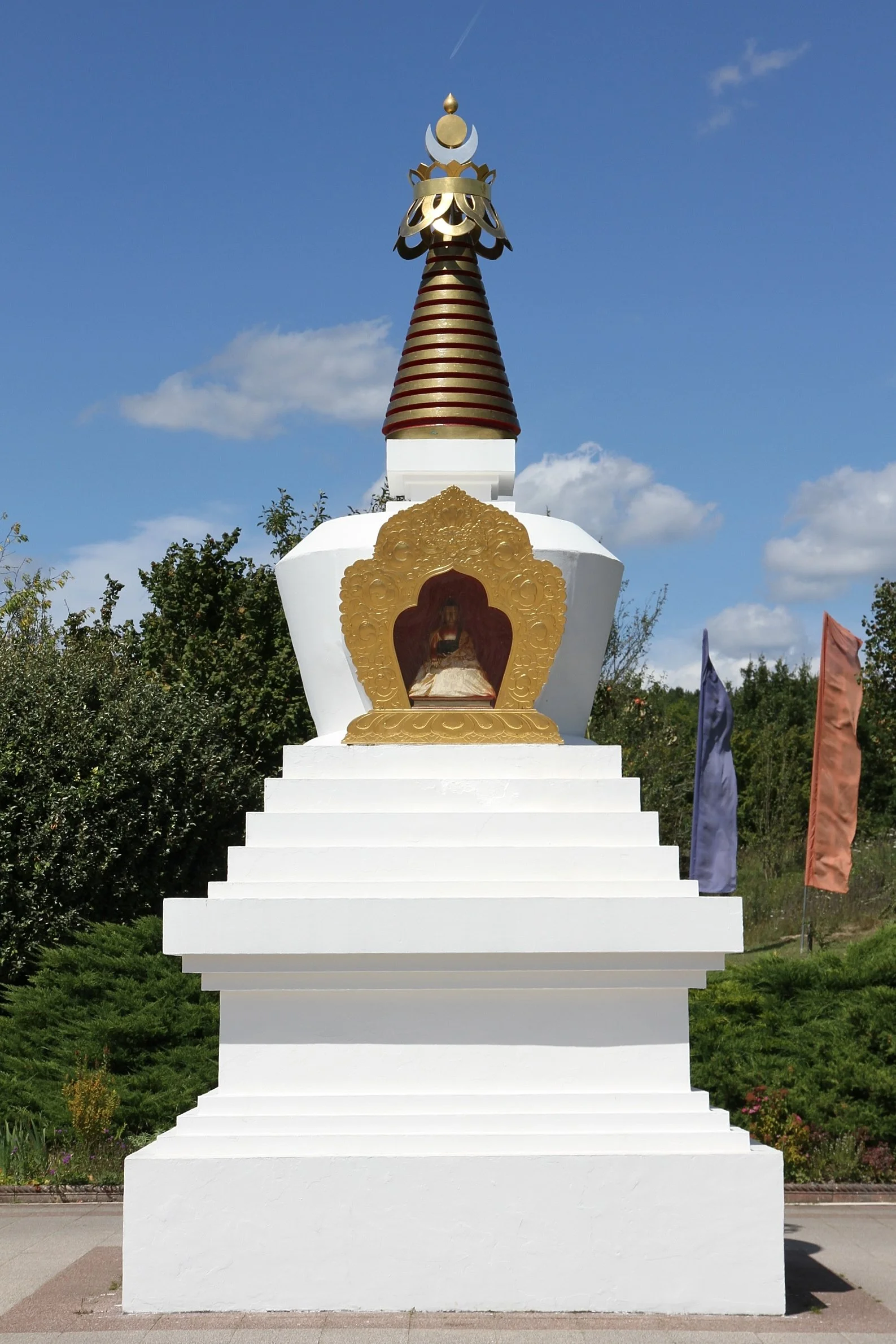 Colorful Buddhist stupa with a small Buddha statue inside a golden frame, set against a blue sky with clouds, surrounded by green trees and flags.
