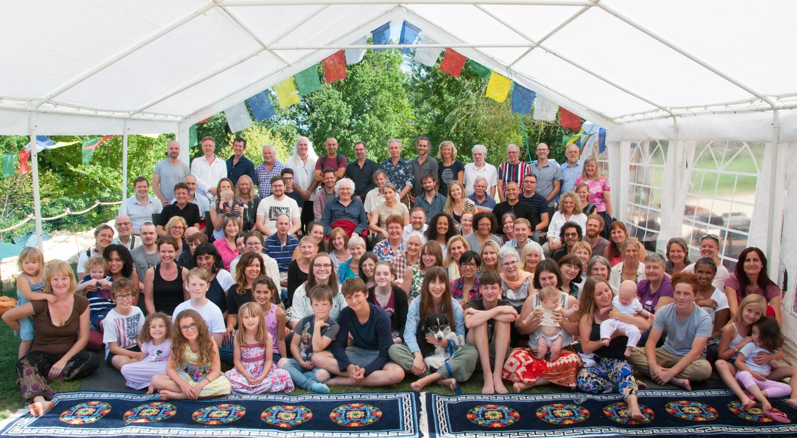 A large group of people, including children, adults, and elderly, gathered inside a decorated white tent outdoors, with trees and greenery visible in the background.