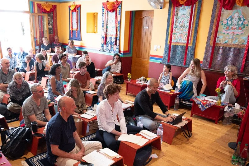 Group of people attending a seminar or workshop in a colorful room with Buddhist art tapestries on the walls.