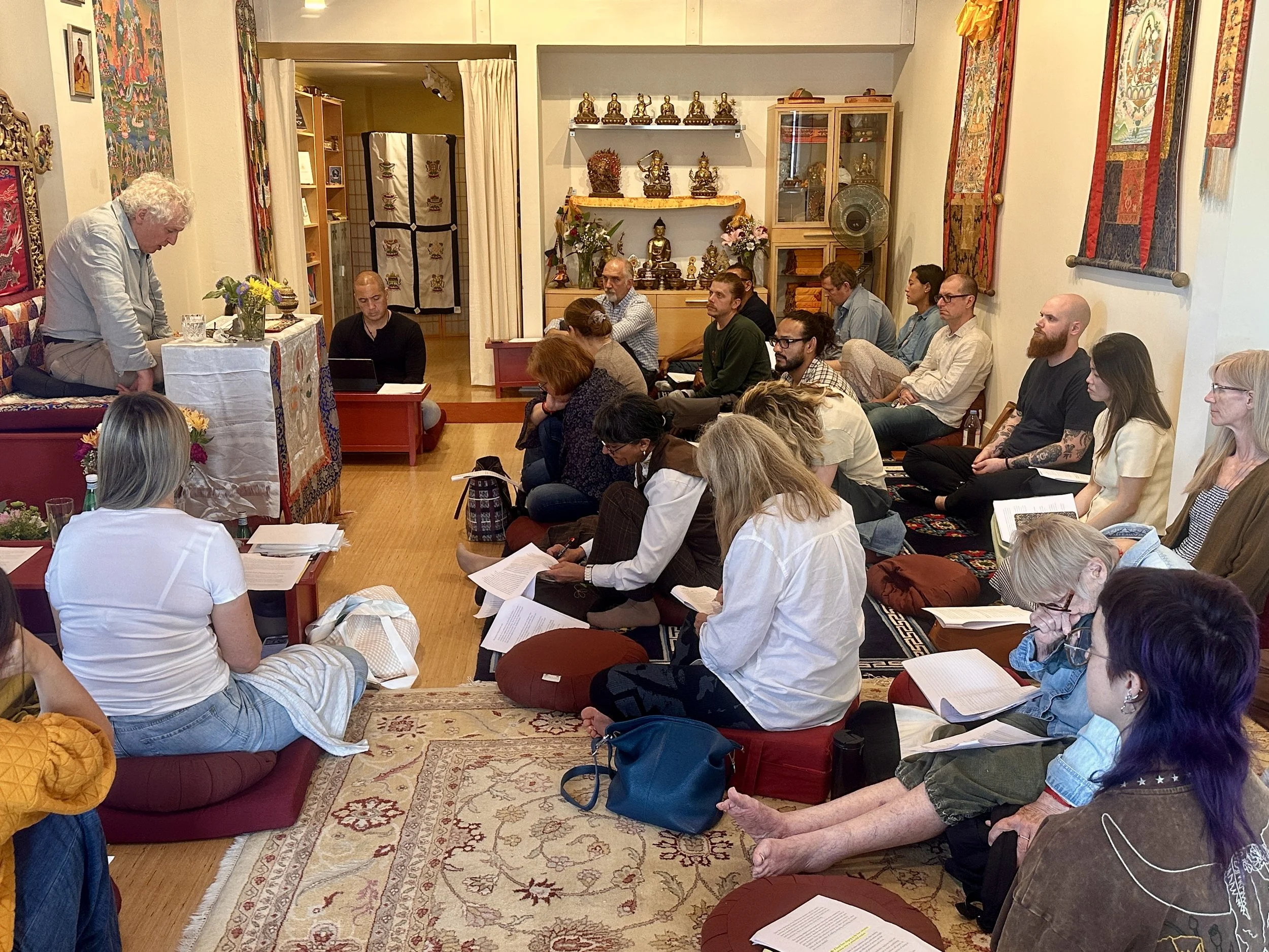 A group of people attending a spiritual or meditation session in a cozy room with Asian artwork and Buddha statues, with a person leading the session at a small altar.