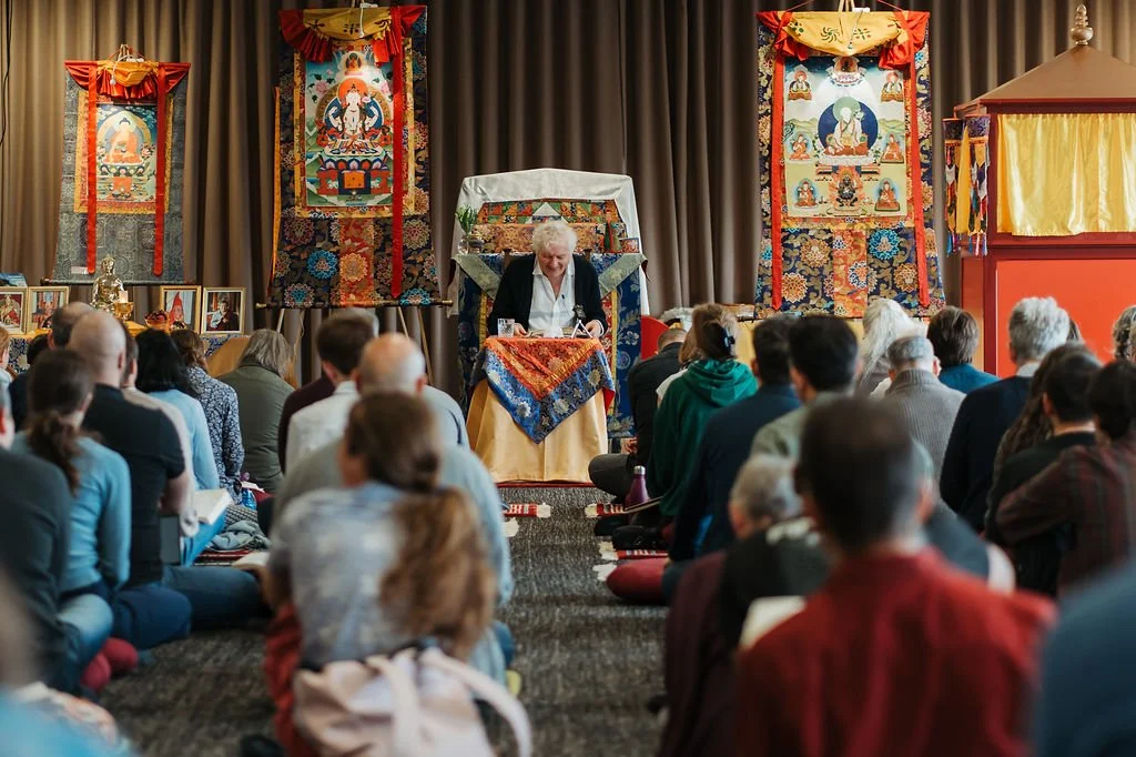 A woman with white hair speaking at a podium with Tibetan Buddhist thangka paintings hanging behind her during a gathering or teaching. People seated on the floor are listening attentively.