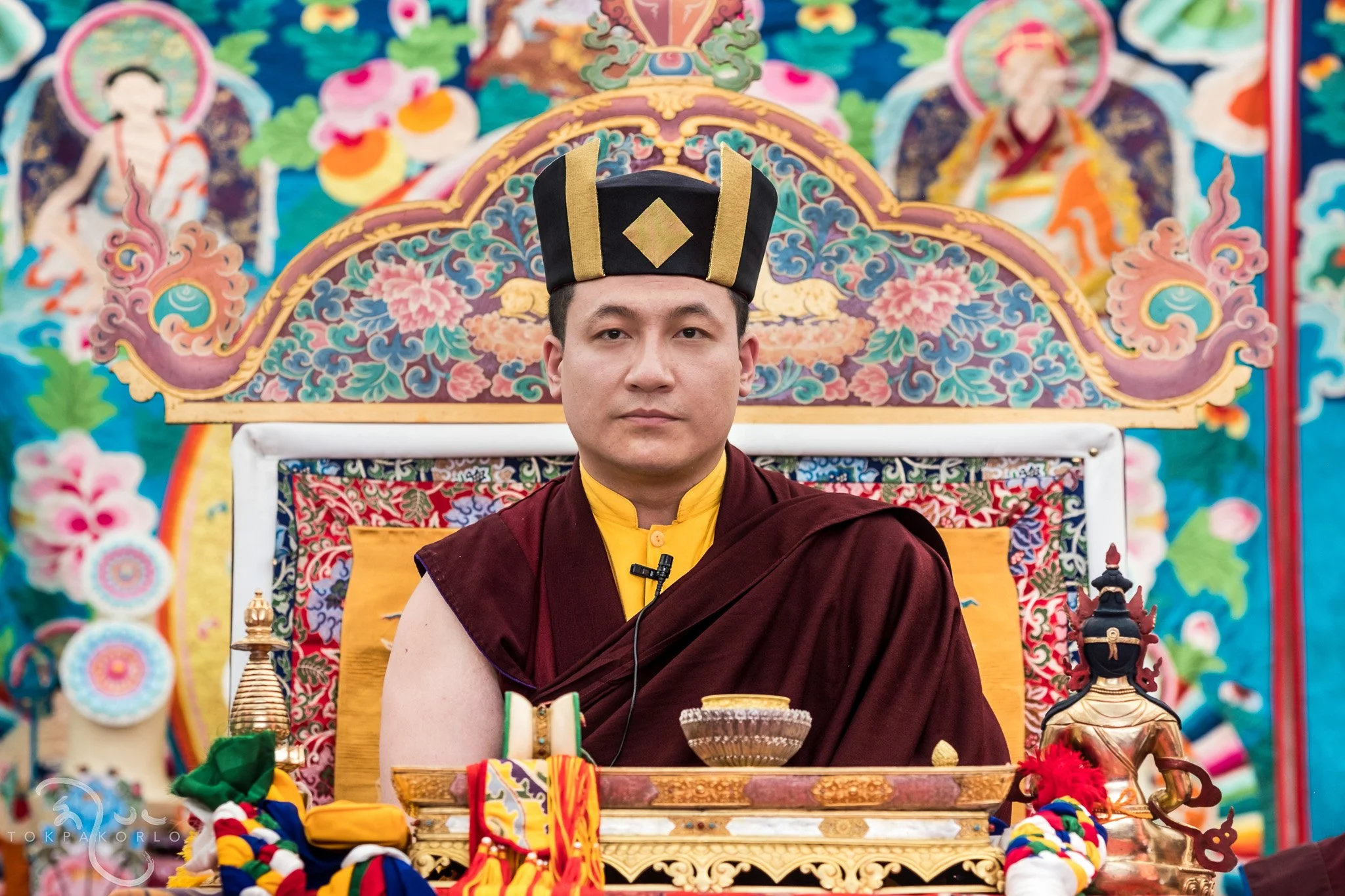 A Buddhist monk seated on an ornately decorated throne in front of a colorful, intricate backdrop featuring religious and traditional motifs. The monk is wearing maroon robes and a black and gold ceremonial hat, with a serious expression.
