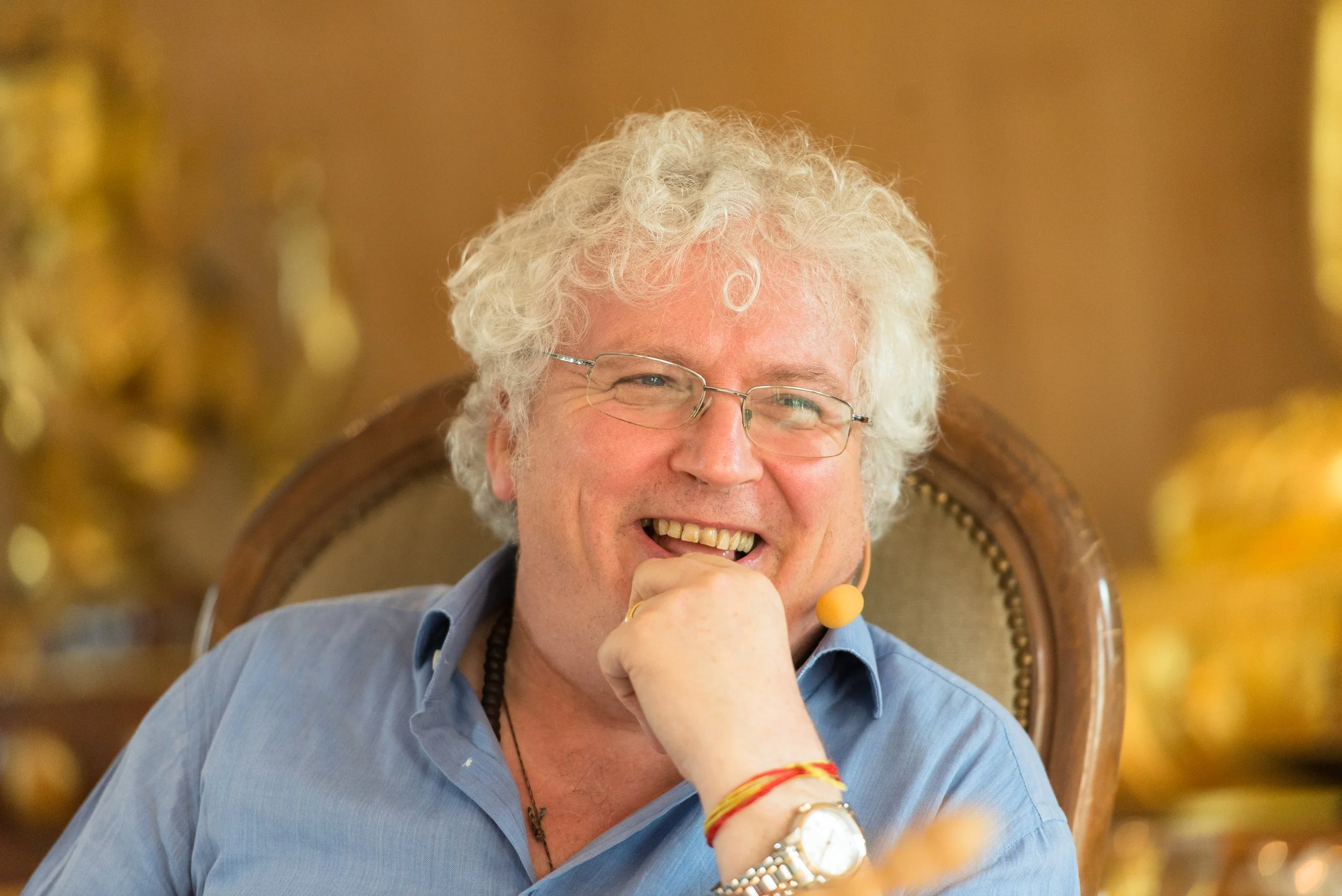 A cheerful man with curly white hair, glasses, wearing a blue shirt, gold watch, red bracelet, and a yellow microphone, sitting on an ornate wooden chair with a high back, smiling and resting his hand on his chin.