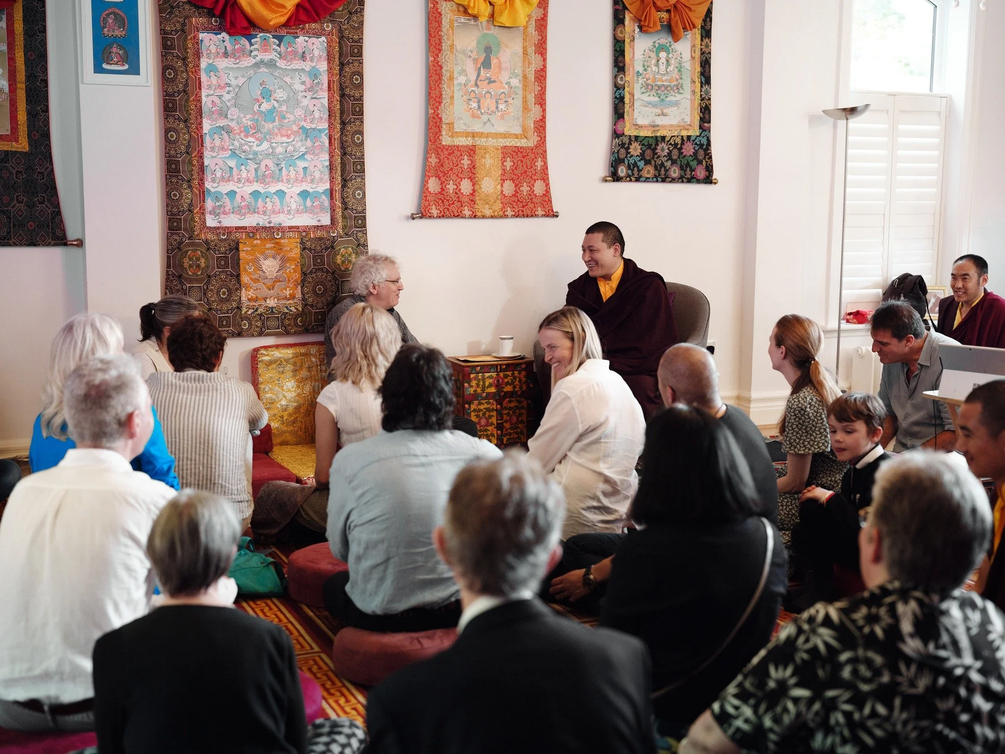Group of people seated on the floor in a meditation class, gathered around a Buddhist teacher with Tibetan thangka paintings on the wall behind him.