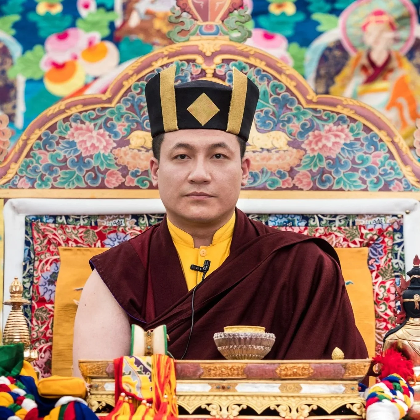 A young monk in traditional maroon and yellow robes sitting at a decorated table, with a colorful ornate backdrop featuring floral patterns and mythical creatures.