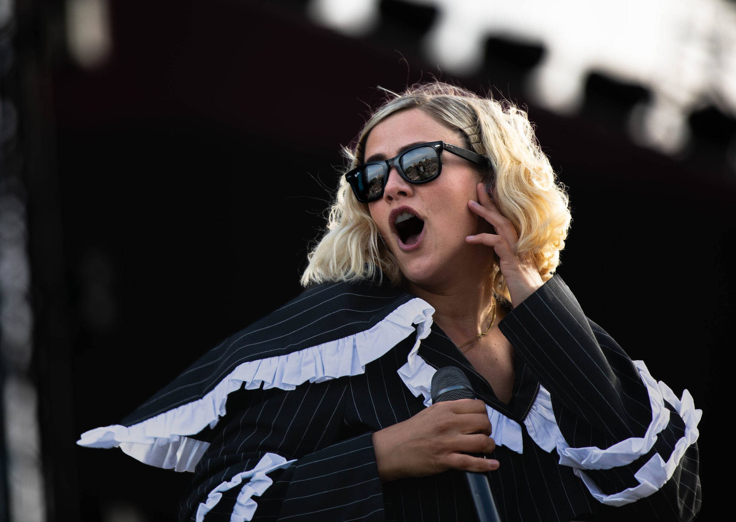 A woman with shoulder-length blonde curly hair wearing black sunglasses and a black and white ruffled jacket, singing into a microphone with her mouth open and hand touching her ear.
