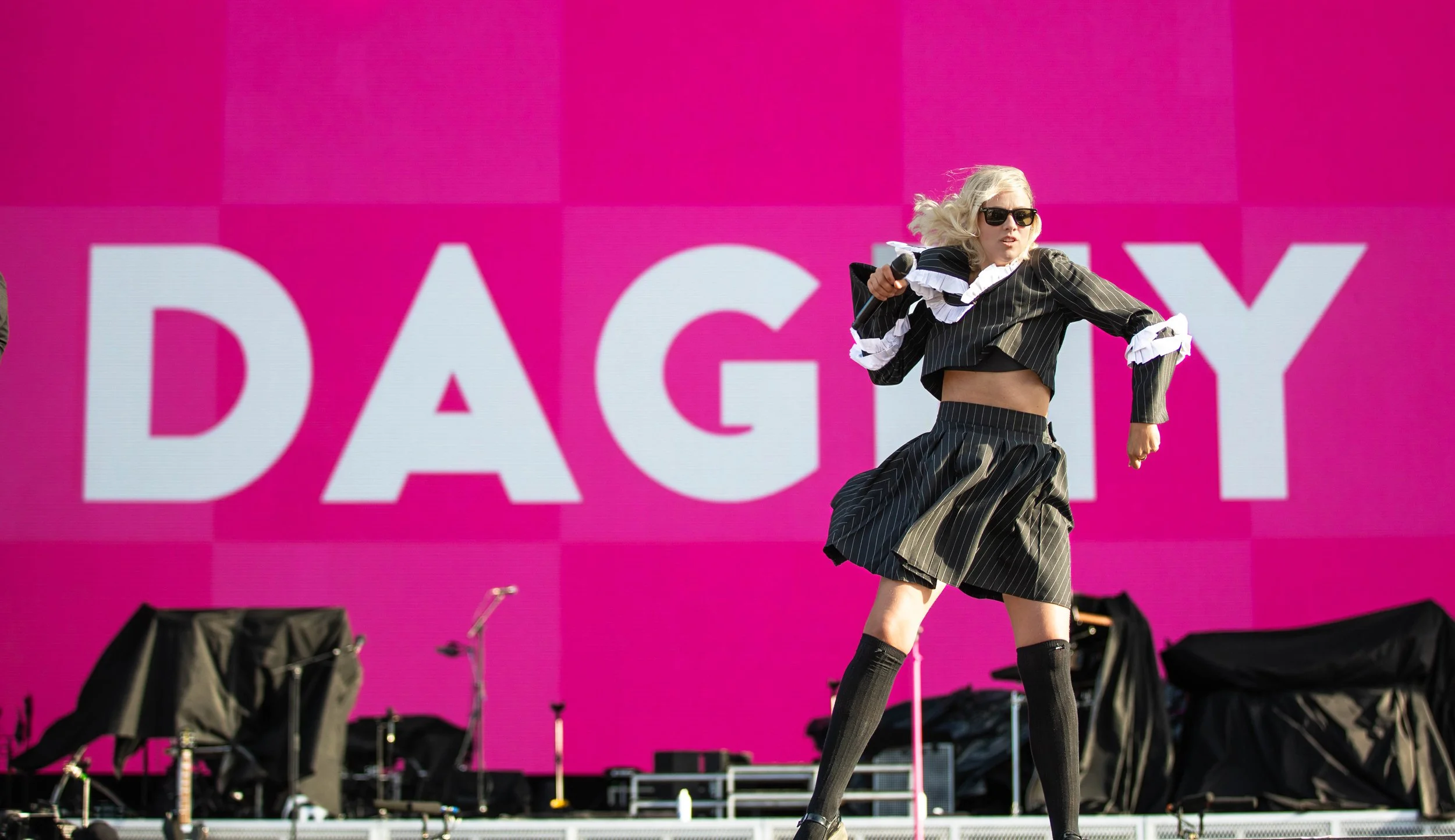 A woman with blonde hair, sunglasses, and a black pinstripe outfit, including a cropped blazer and a skirt, performs on stage during a daytime event in front of a large pink background with white text reading 'DAY'. She holds a microphone and appears