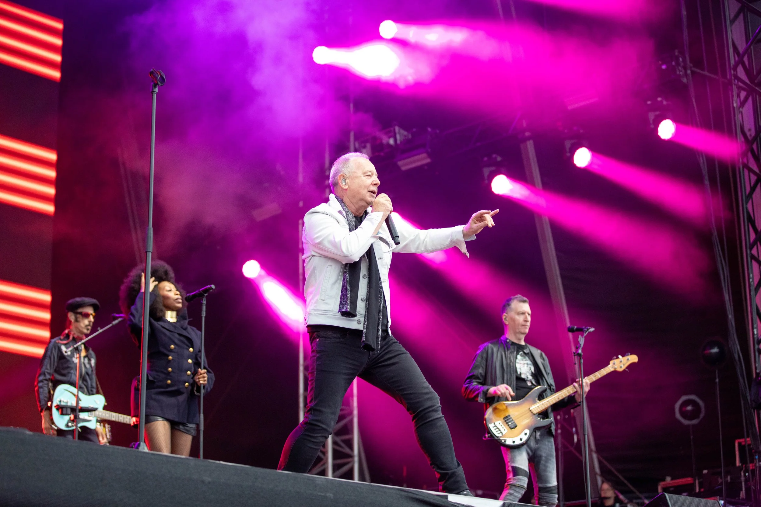 Performers on stage during a concert with purple stage lights, including a man in a white jacket singing and pointing, a woman with an afro singing, and two men playing guitars.