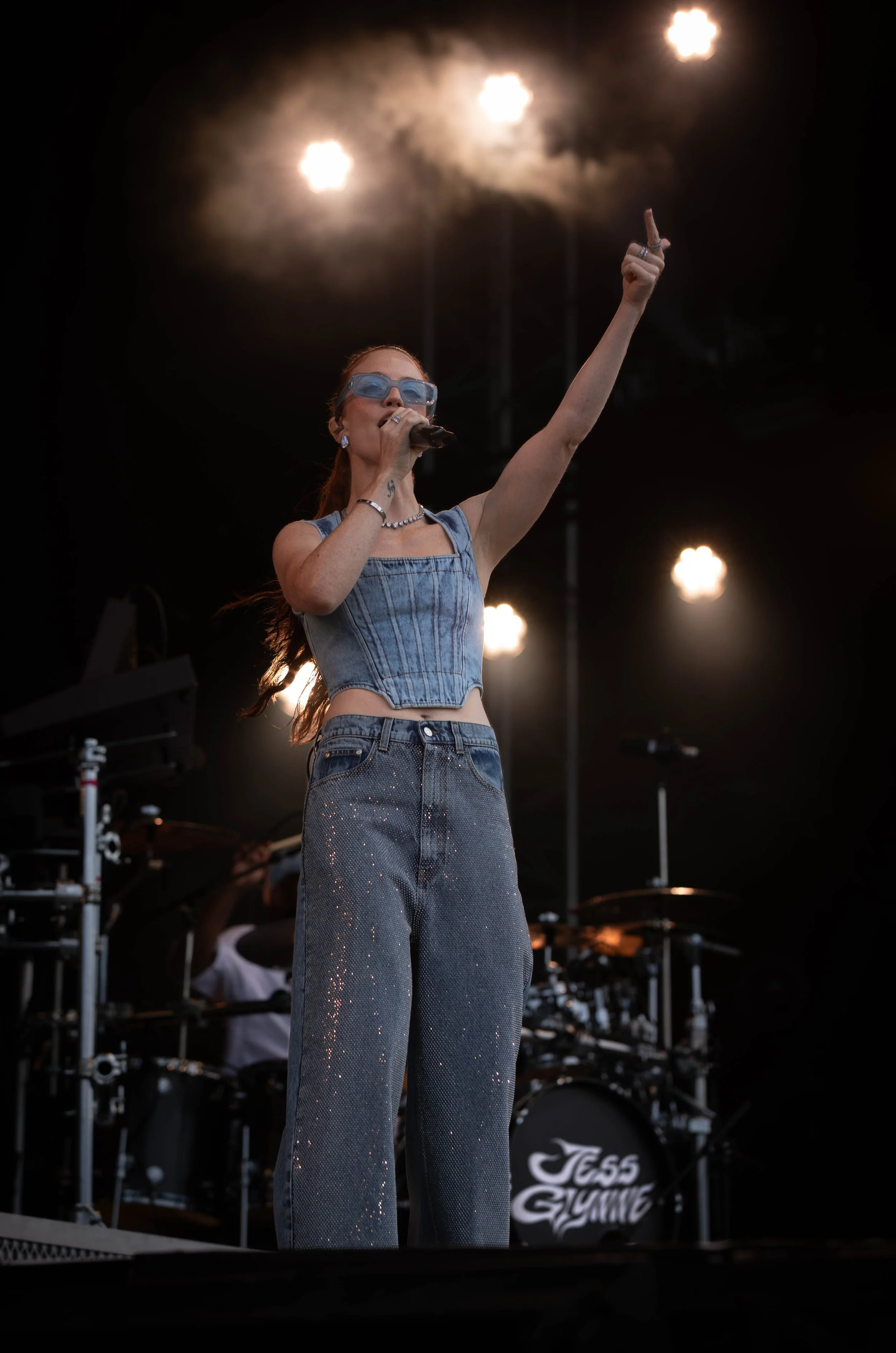 A female singer performing on stage with a microphone, wearing sunglasses, a denim crop top, and glittery jeans, with a musician playing drums in the background.