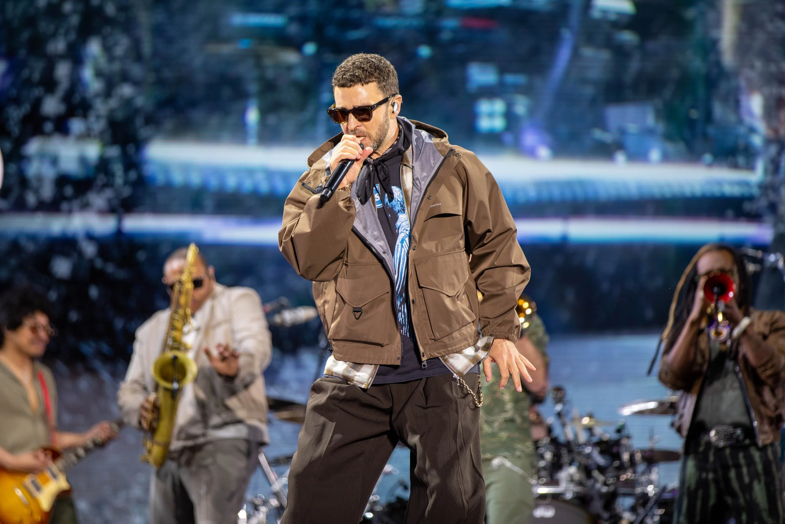 A man with sunglasses singing into a microphone on stage, surrounded by band members playing guitars and brass instruments, with a large screen displaying a scenic background behind them.