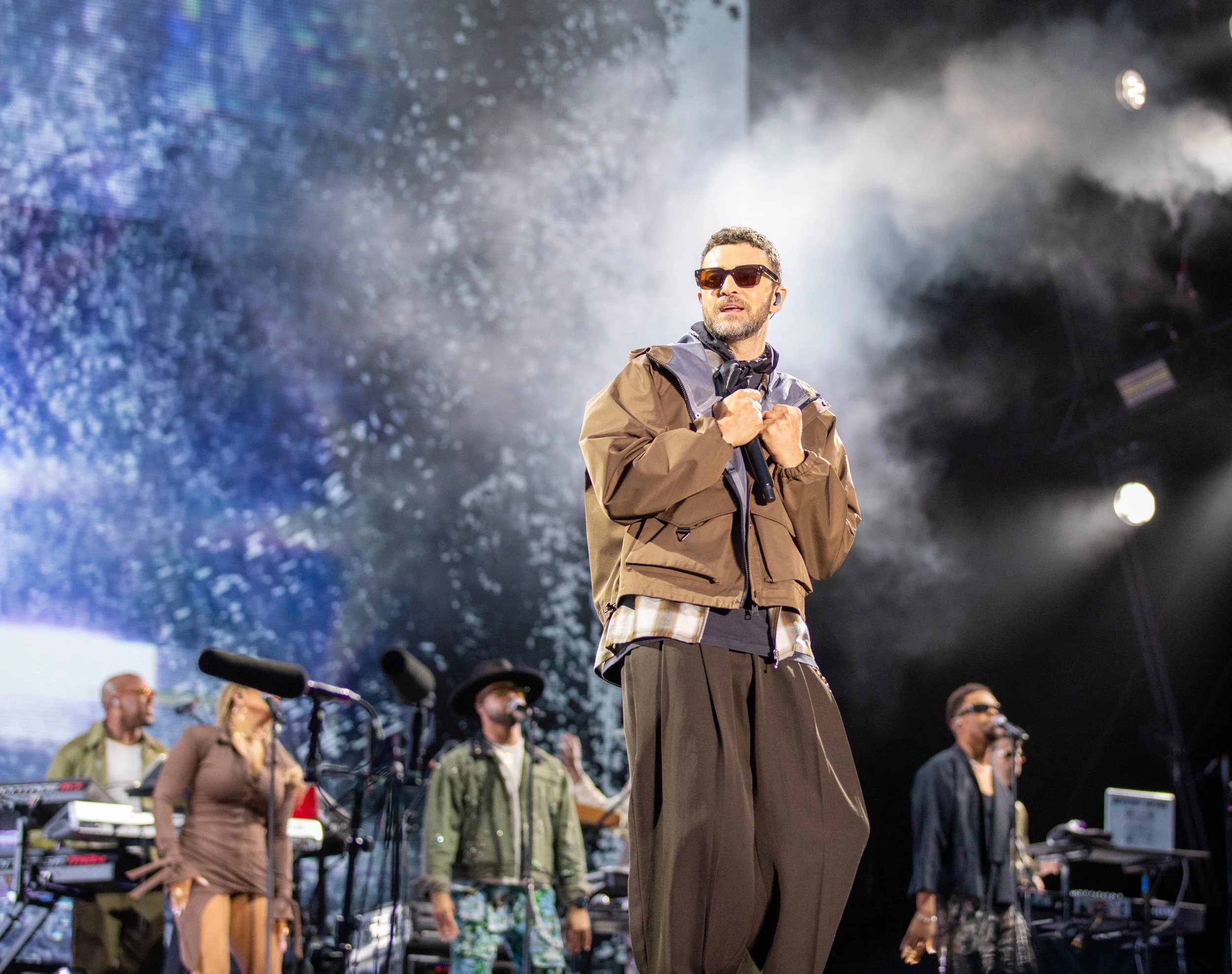 A man in sunglasses, a brown jacket, and baggy pants holds a microphone on stage during a concert with a group of performers and a large screen showing water or snow in the background.