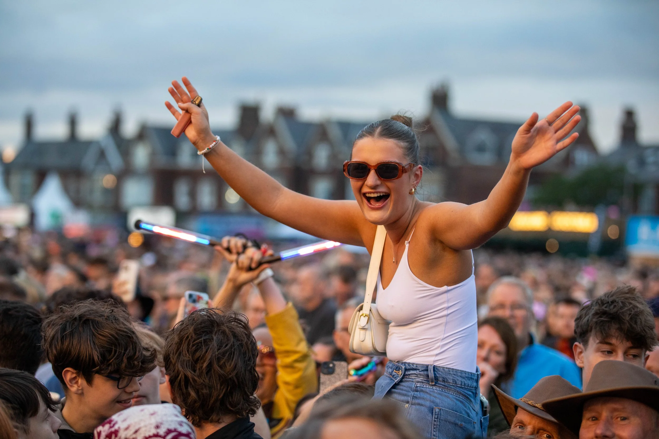 A young woman in sunglasses and white tank top celebrating in a crowd, raising her arms with a smile, during an outdoor festival.