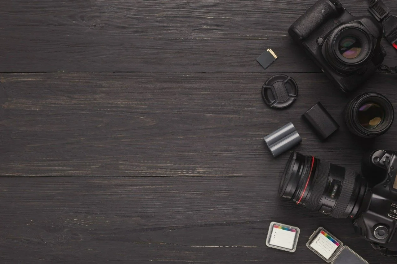 Photography equipment including camera bodies, lenses, memory cards, and a camera strap on a dark wooden surface.