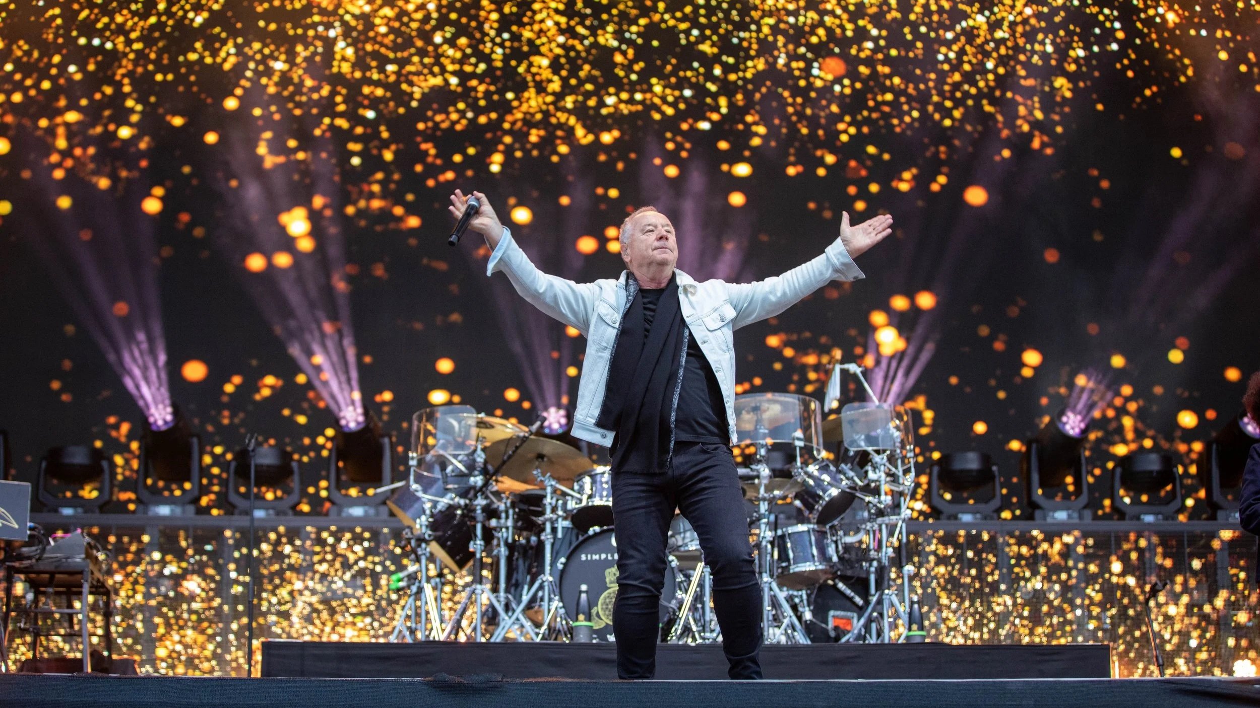 Performer on stage with arms outstretched, holding a microphone, against a backdrop of colorful lights resembling glowing orbs.