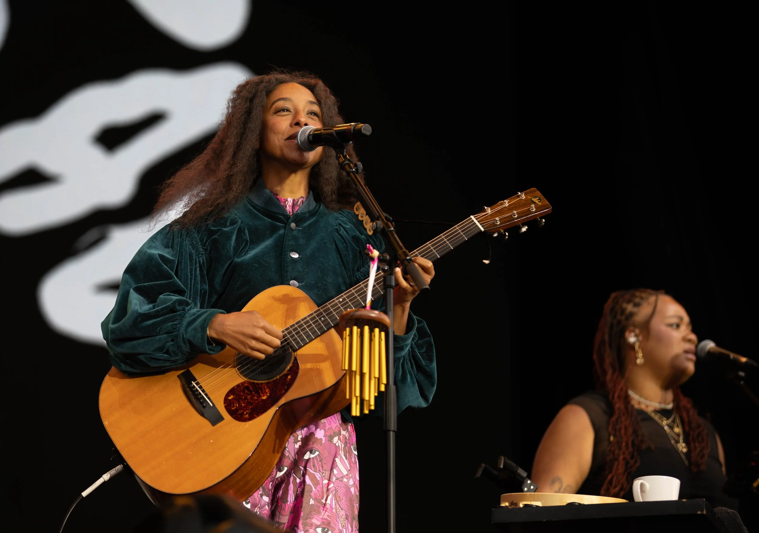 Two women perform on stage, one playing an acoustic guitar and singing into a microphone, the other at a keyboard with a teacup. Both women have long braided hair, and the background features a large abstract black and white design.