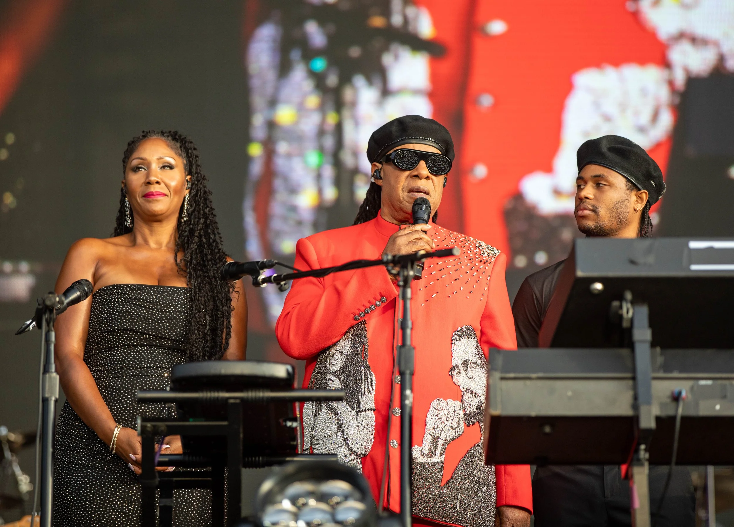 Three performers standing on stage during a musical event. The woman on the left is wearing a black dress with white polka dots and has long, twisted hair. The man in the middle is wearing a bright red jacket with silver embellishments, a black hat, 