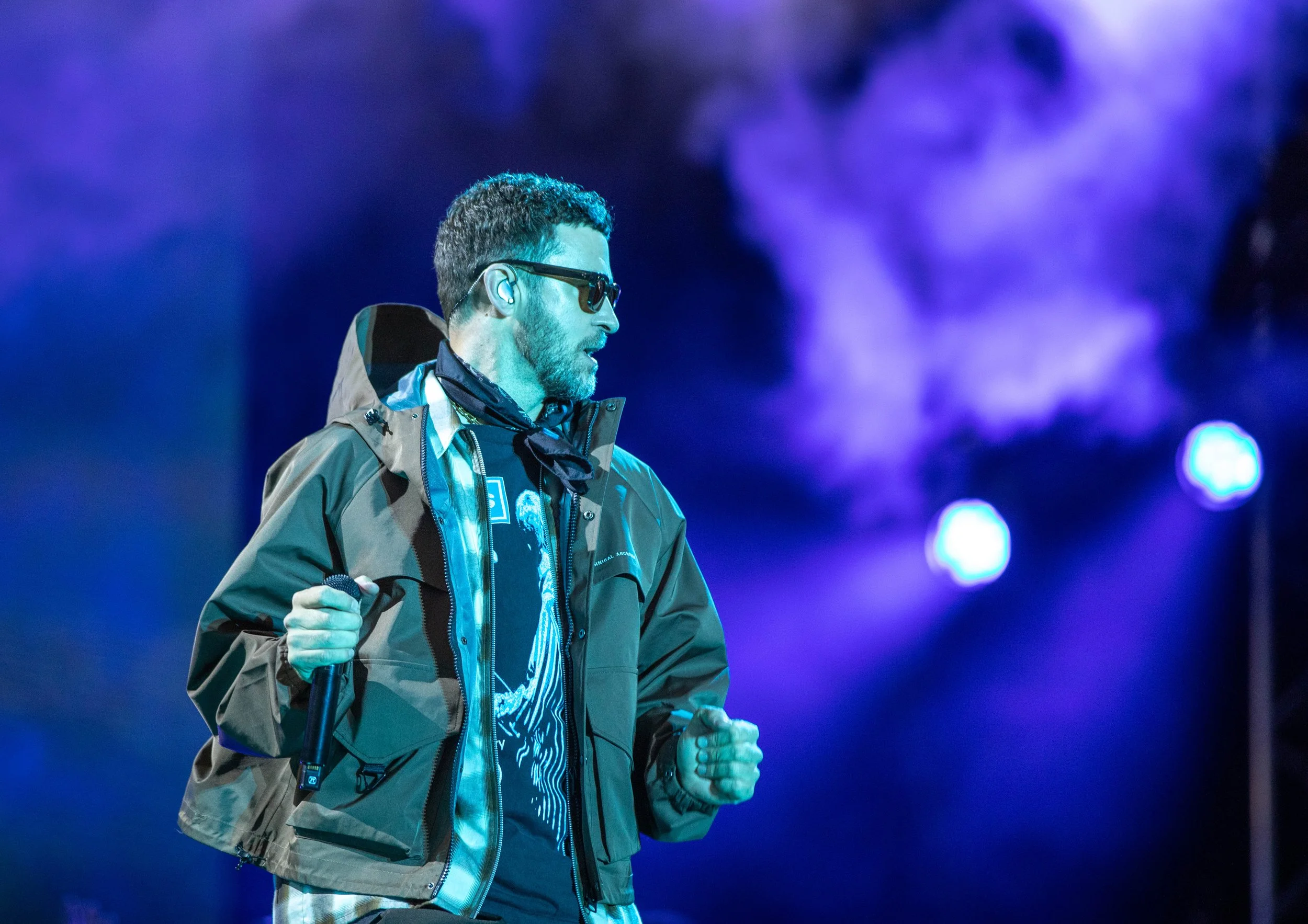 A man wearing sunglasses, a khaki outdoor jacket, and a graphic t-shirt, holding a microphone, stands on a stage with purple lights and fog in the background.