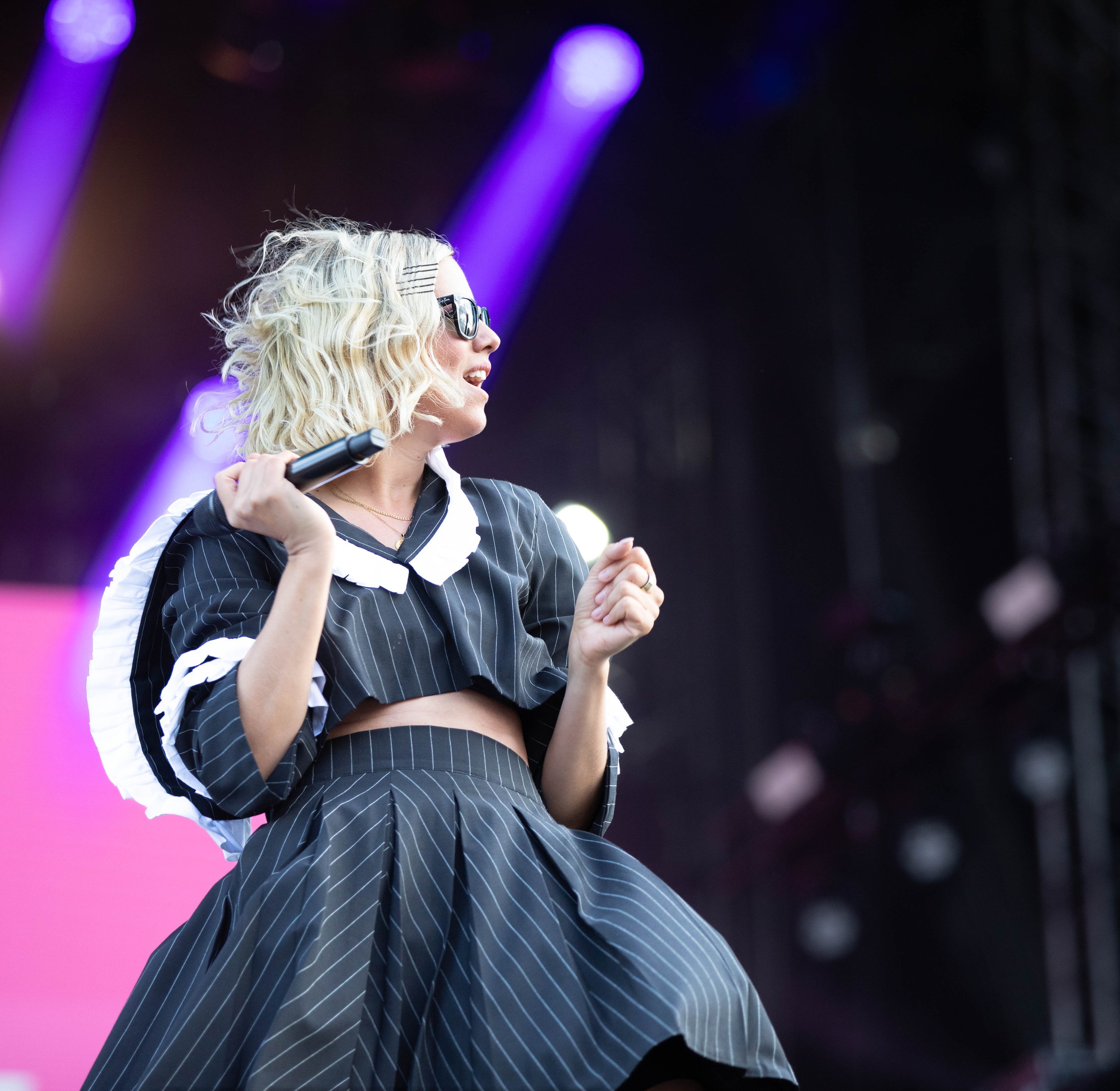 A woman with curly blonde hair, wearing sunglasses and a black pinstripe outfit with white accents, is holding a microphone on a stage with purple lighting.