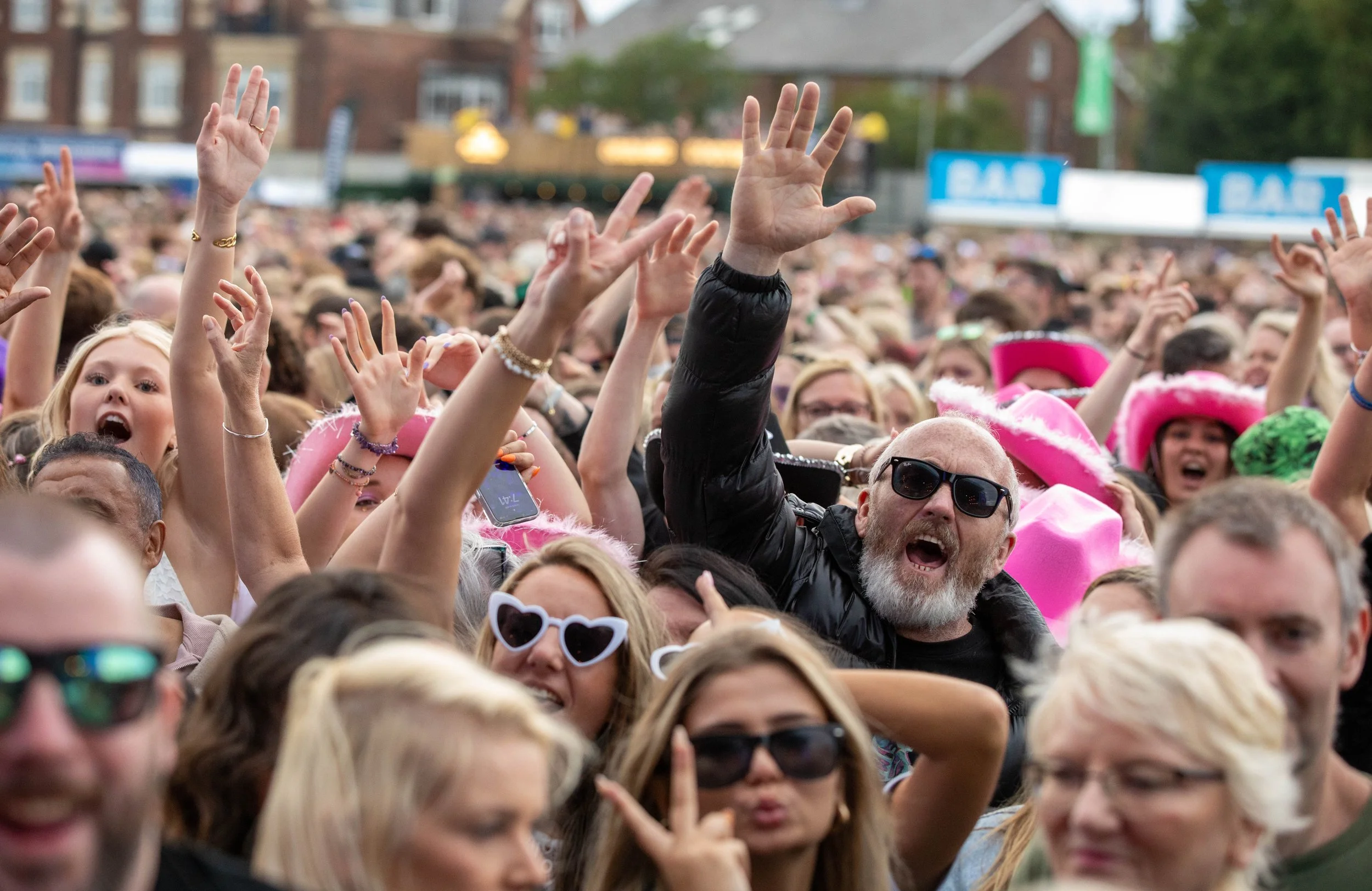 Crowd of people at an outdoor event, many with hands raised, some wearing sunglasses and pink hats, feeling excited and lively.