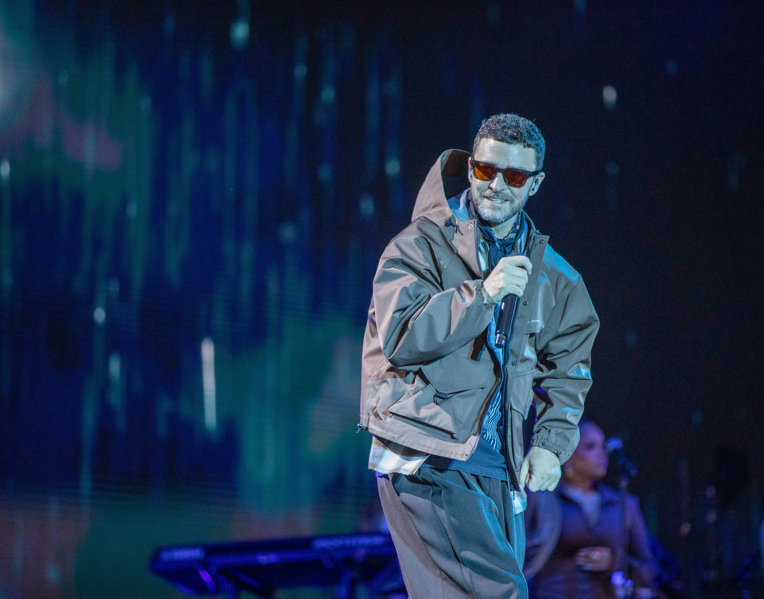 A man wearing sunglasses and a brown jacket singing into a microphone on stage with colorful lights in the background.