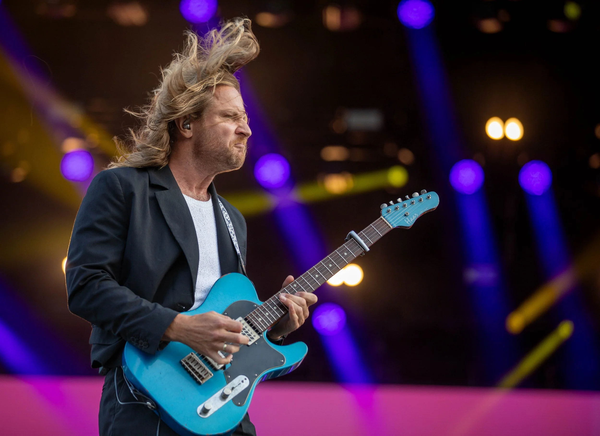 A male guitarist with long, blond hair playing an electric blue guitar on stage with colorful lights in the background.
