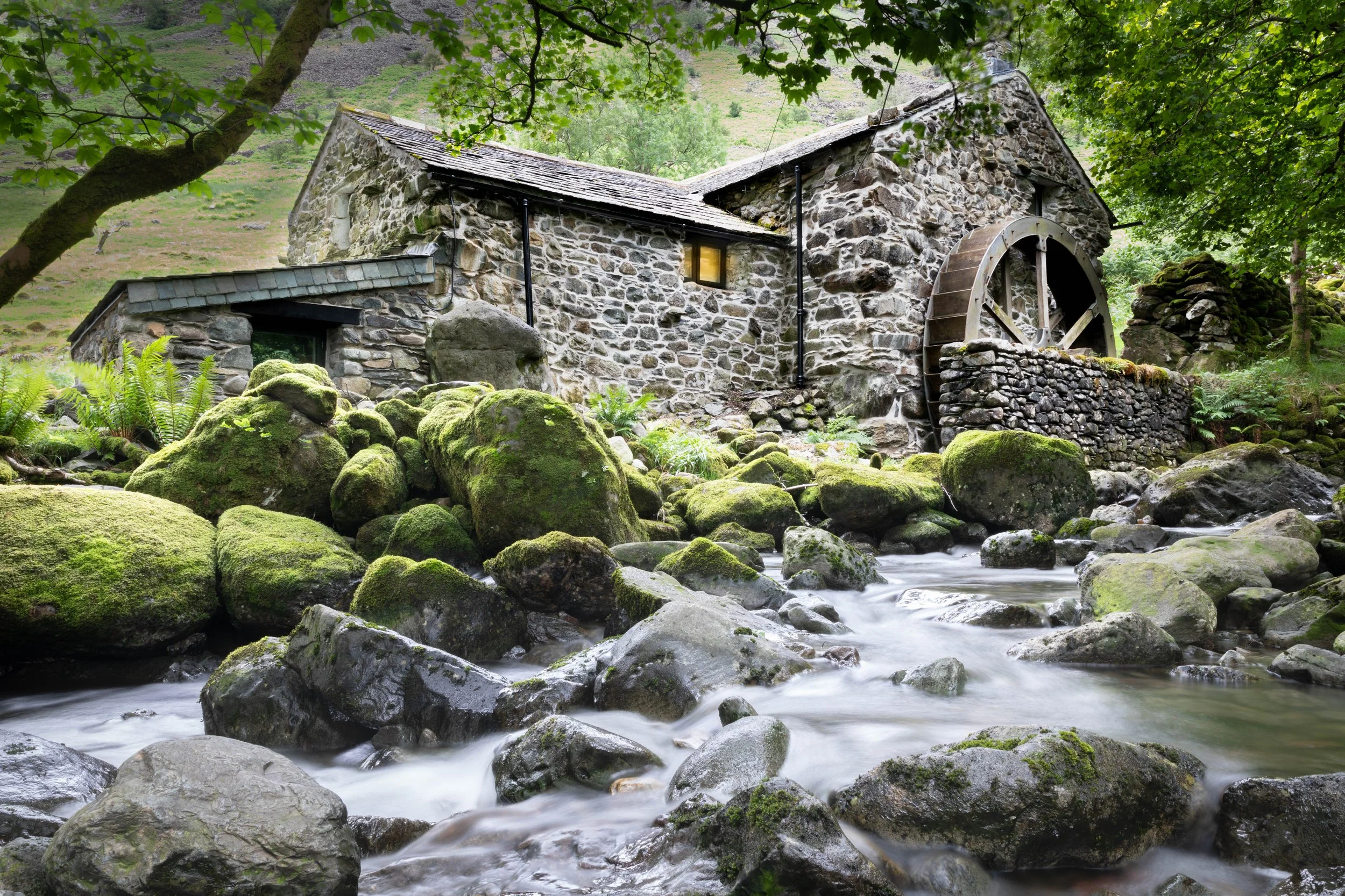 An old stone watermill with a wooden water wheel next to a flowing stream surrounded by moss-covered rocks and greenery in a lush forest setting.