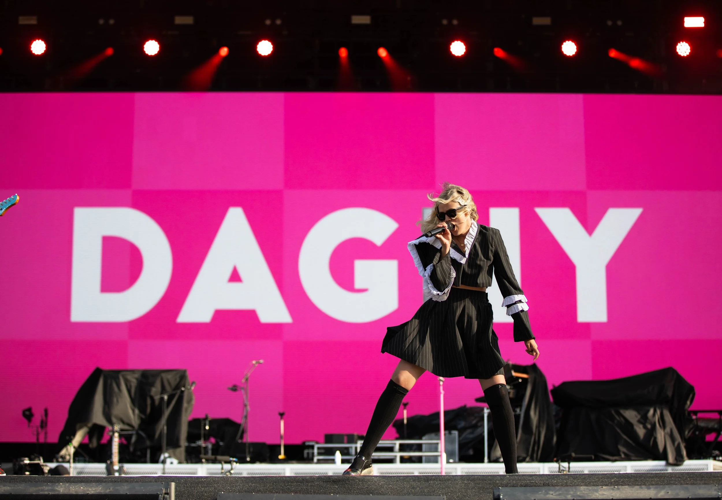 Performer singing on stage with a large pink screen behind showing the word 'DAGY' in white letters, dressed in a black pinstripe suit, thigh-high socks, and sunglasses.