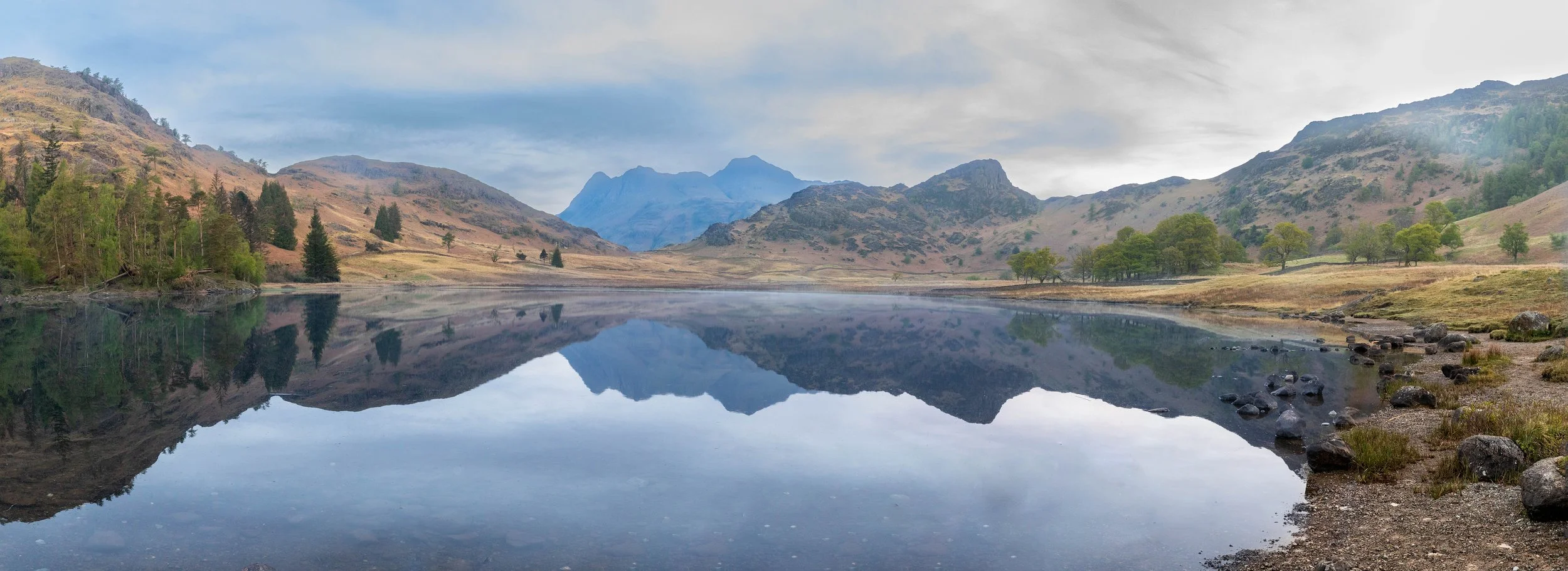Mountain landscape with a calm lake reflecting the mountains, trees, and sky, with rocky shoreline in the foreground.