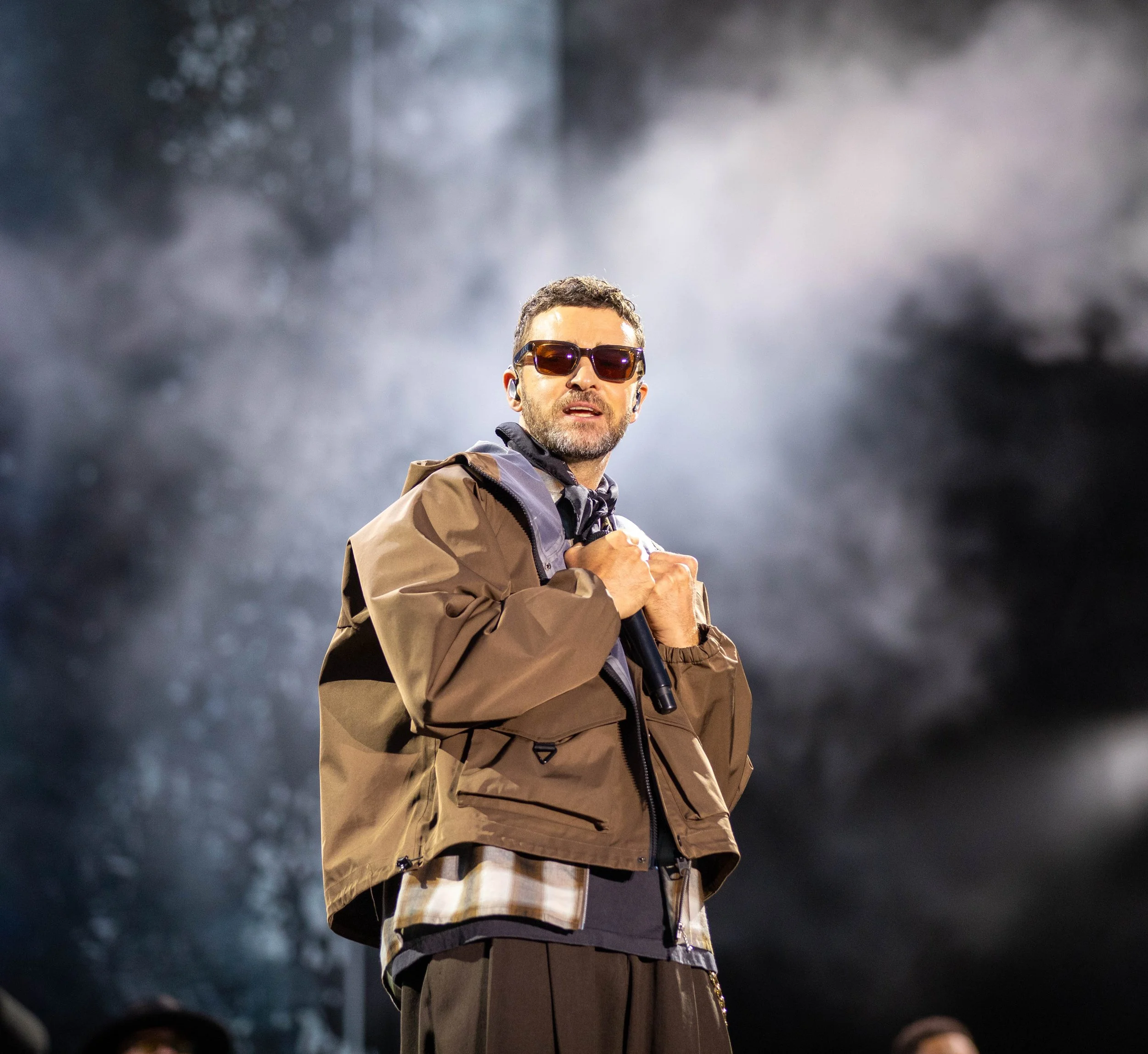 A man wearing sunglasses and a brown jacket holding a microphone on stage with smoke and lights in the background.