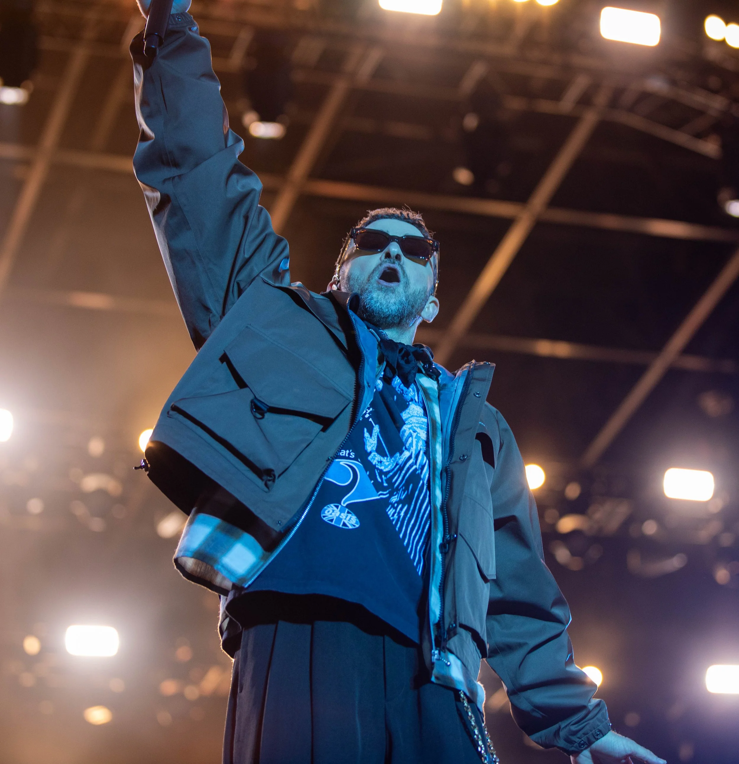 Man performing on stage with sunglasses, wearing a black shirt, jacket, and pants, with stage lights overhead.
