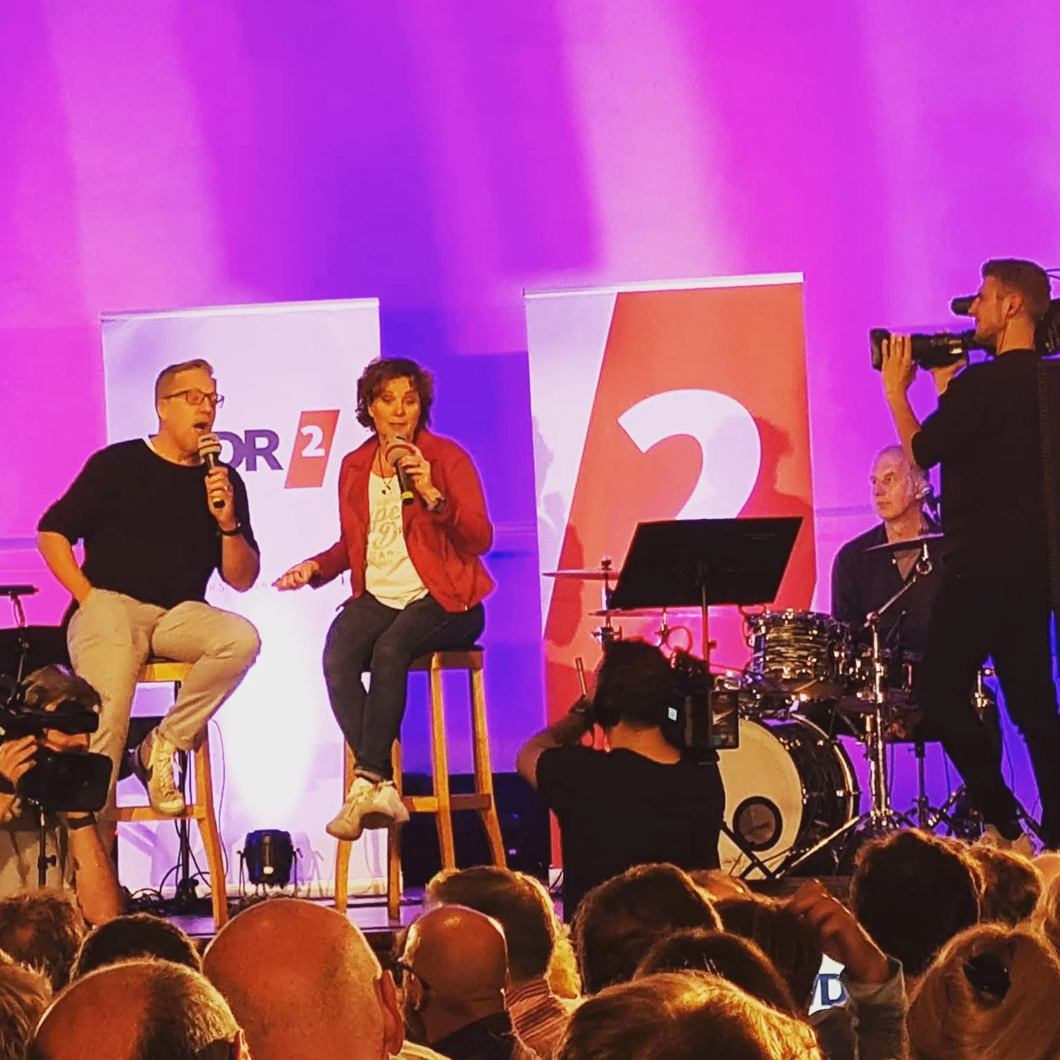 Zwei Personen sitzen auf Barhockern auf einer Bühne und unterhalten sich, während ein dritter Mann mit einer Kamera filmt. Es gibt musikalische Instrumente im Hintergrund und ein Publikum im Vordergrund. Es sind Banner mit den Logos von ARD1 und 2 si