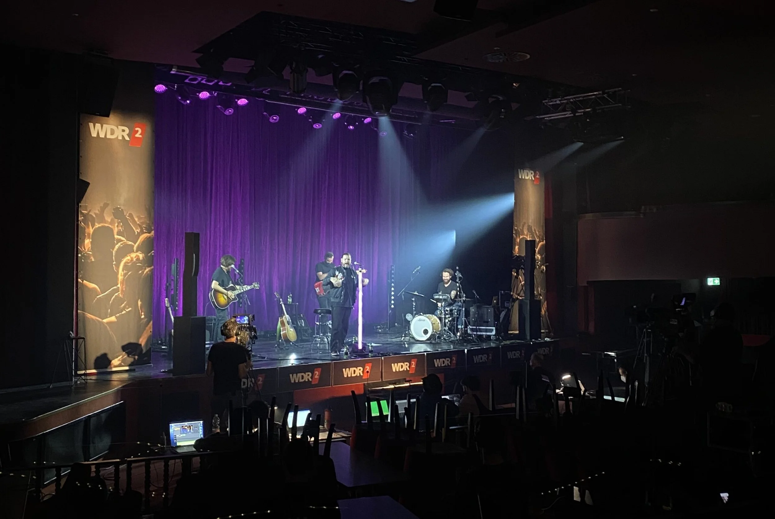 Musiker auf einer Bühne bei einem Live-Konzert, mit mehreren Instrumenten und Beleuchtung, im Hintergrund ein purple Vorhang, Branding von WDR 2 sichtbar.