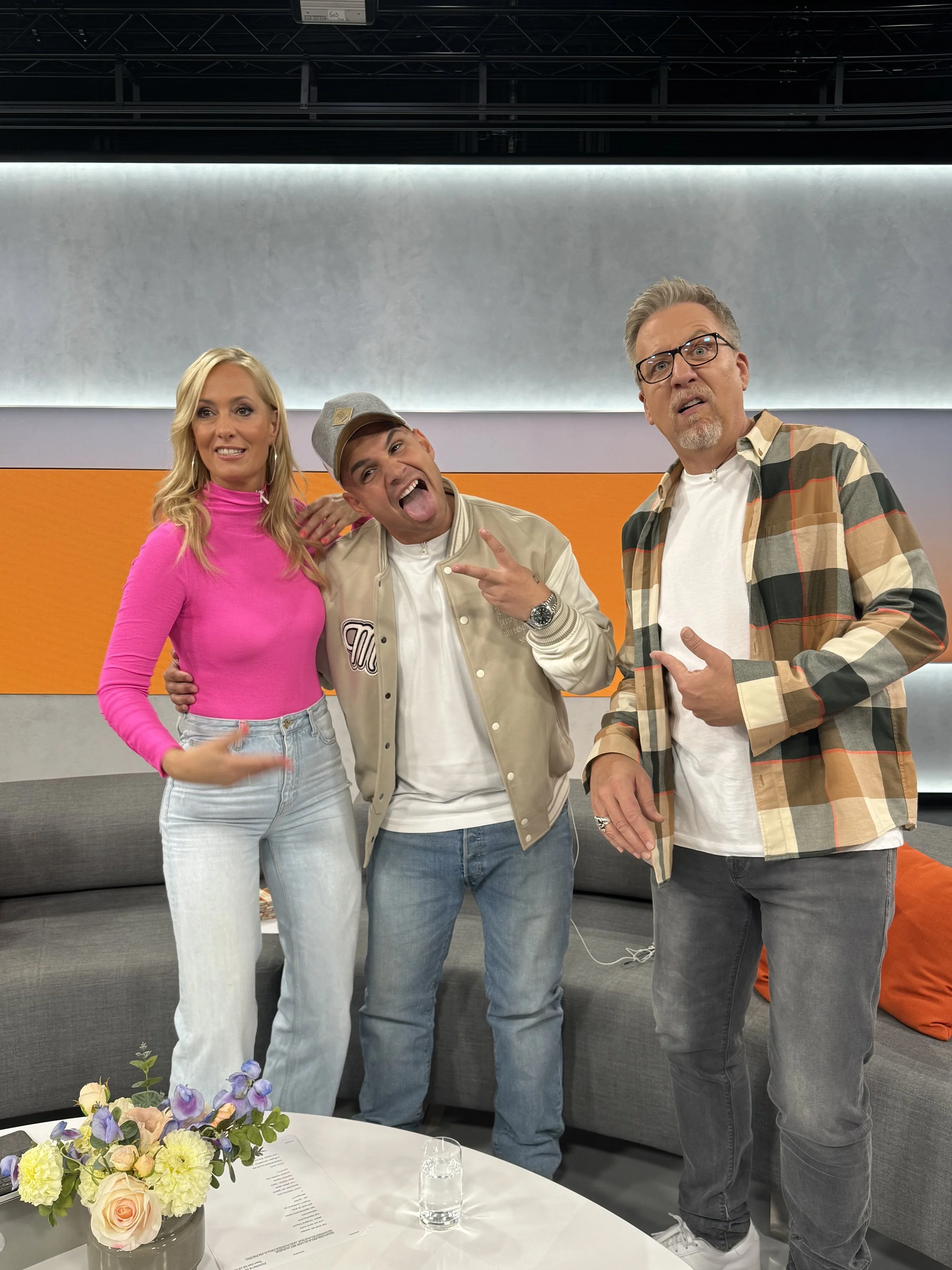 Drei Personen posieren lachend im Studio. Ein Mann trägt einen grauen Baseballmantel, ein Mann trägt eine kariert Jacke und eine Frau trägt ein pinkes Oberteil. Im Vordergrund steht ein Tisch mit einem Blumenstrauß und einem Glas Wasser.