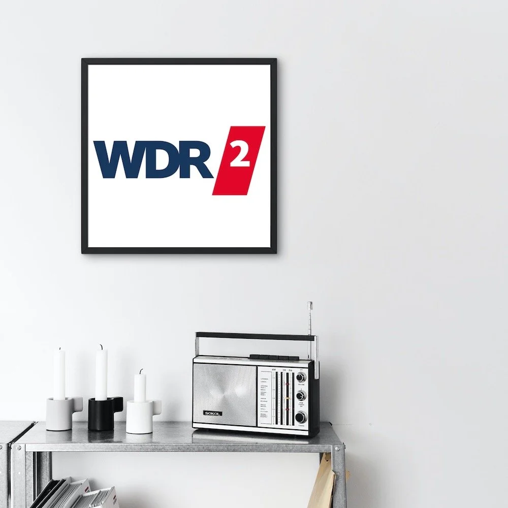 Ein Wandbild mit dem Logo von WDR 2, einem Radiosender, hängt an einer weißen Wand. Darunter steht ein alter schwarzer und silberner Radio auf einem grauen Tisch, begleitet von drei Kerzen in weißen und schwarzen Kerzenhaltern.