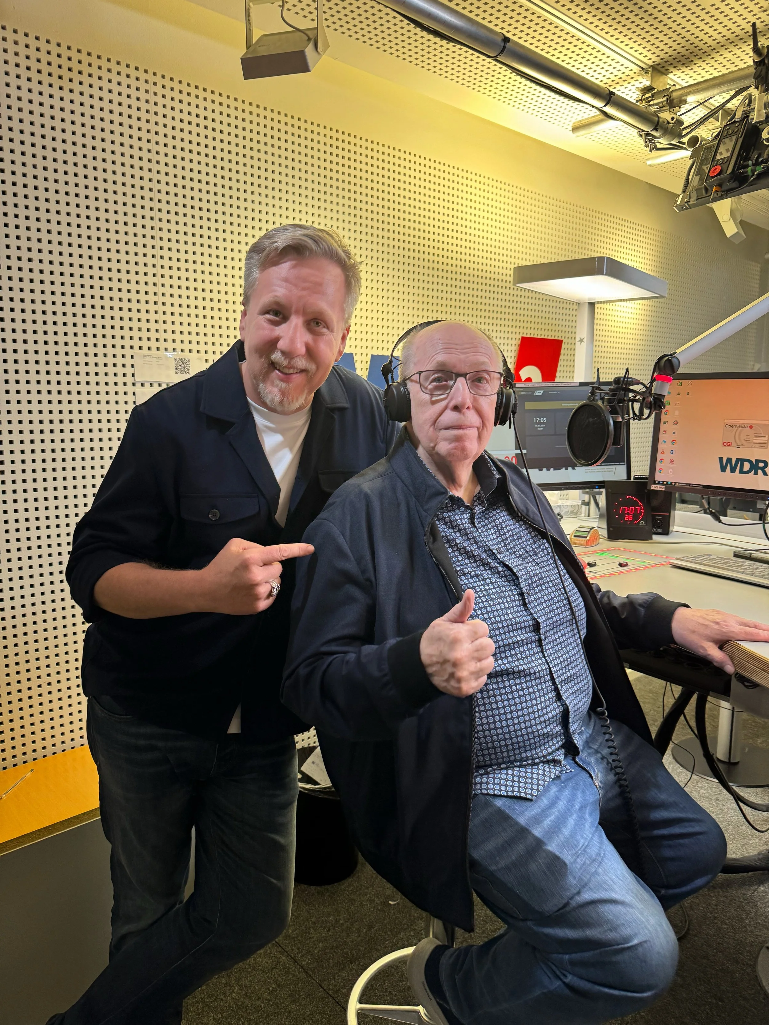 Zwei Männer in einem Radiostudio, einer steht und zeigt auf den anderen, der sitzt und trägt Kopfhörer, umgeben von Bildschirmen und Aufnahmetechnik.