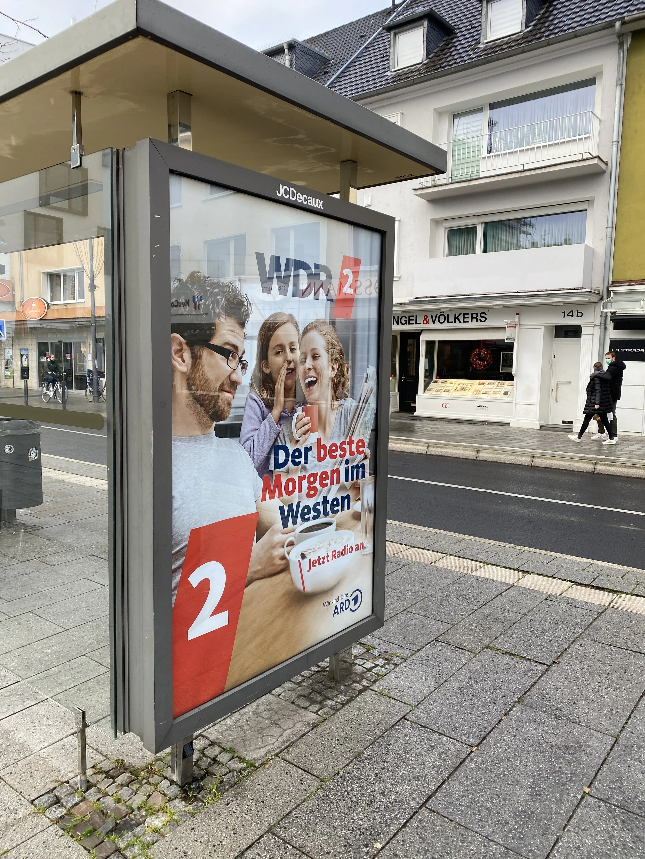 Werbeanzeige für Radio ARD mit einem Mann und zwei jungen Mädchen, die Kaffee trinken und sprechen, auf einer Bushaltestelle in Deutschland.