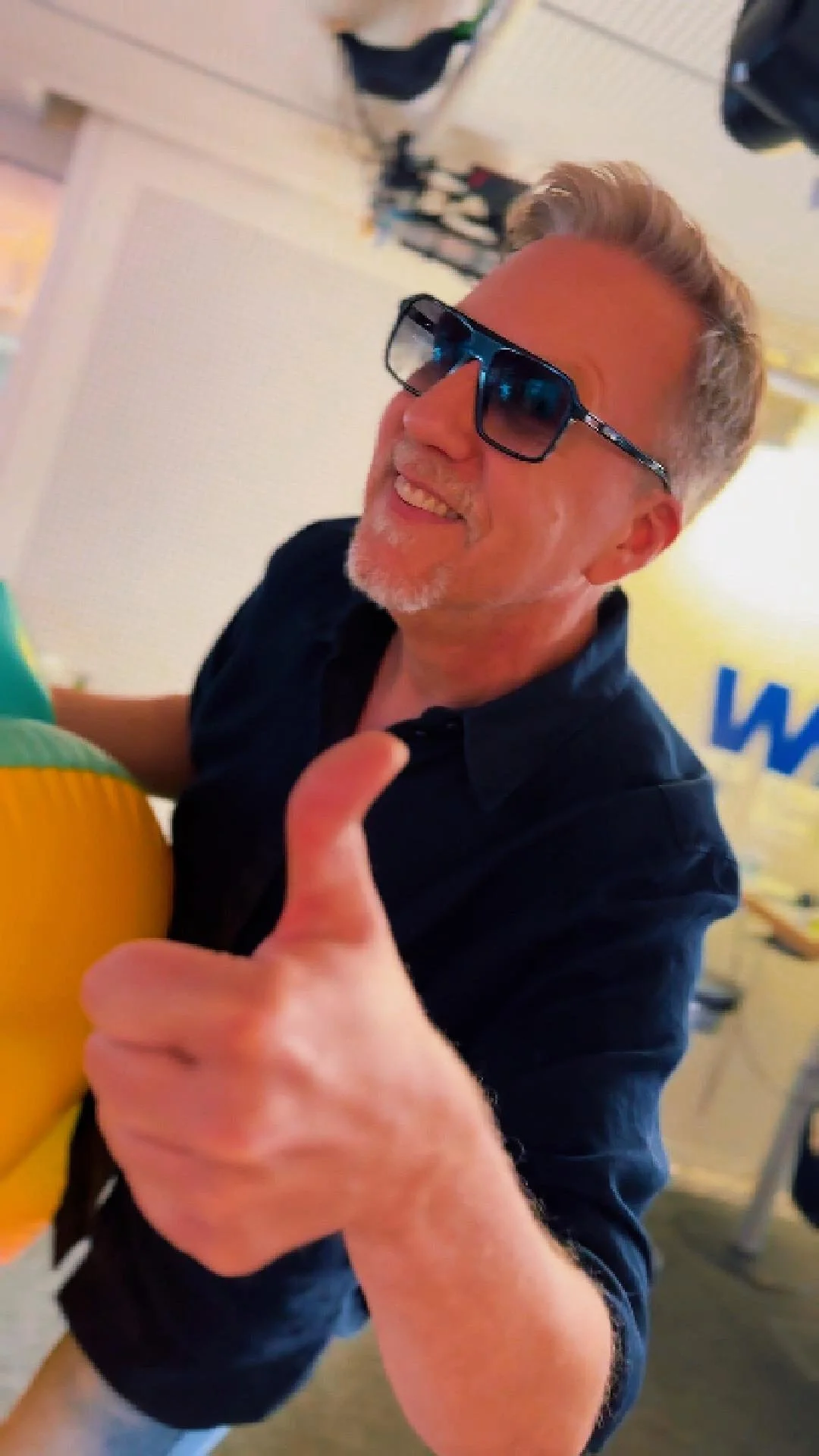 Jan Malte Andresen trägt dunkle Sonnenbrille, zeigt einen Daumen nach oben und ist in einer bunten, lebhaften Umgebung.