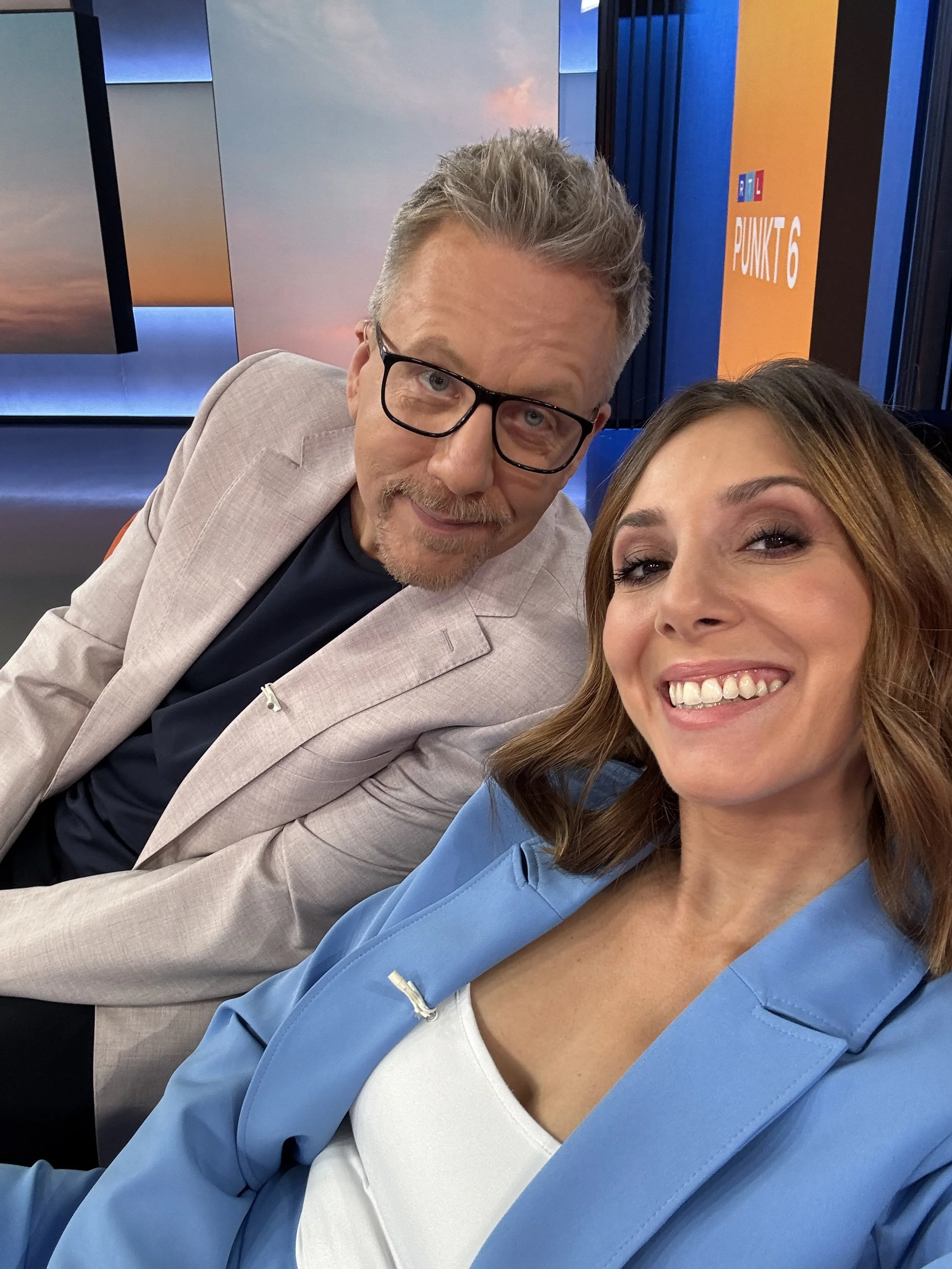 Zwei Fernsehmoderatoren im Studio, beide lächeln und nehmen einen Selfie auf, im Hintergrund ein Bildschirm mit blauer und orangefarbener Gestaltung.