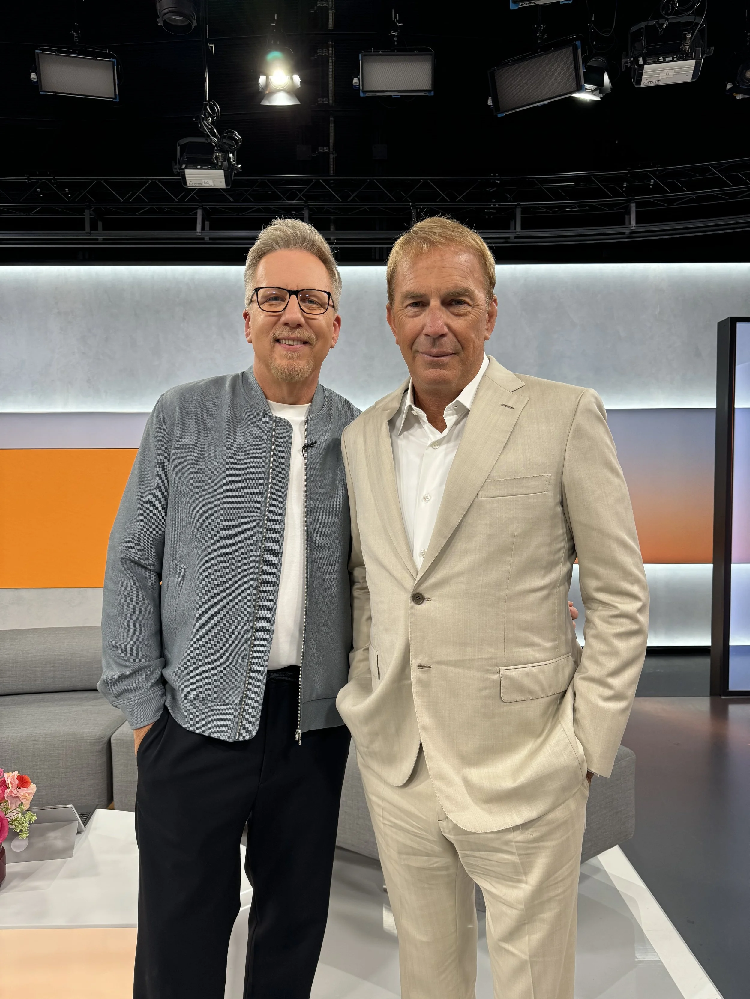 Zwei Männer stehen nebeneinander in einem Fernsehstudio, eines trägt einen grauen Blouson und das andere einen beigefarbenen Anzug, beide schauen in die Kamera.