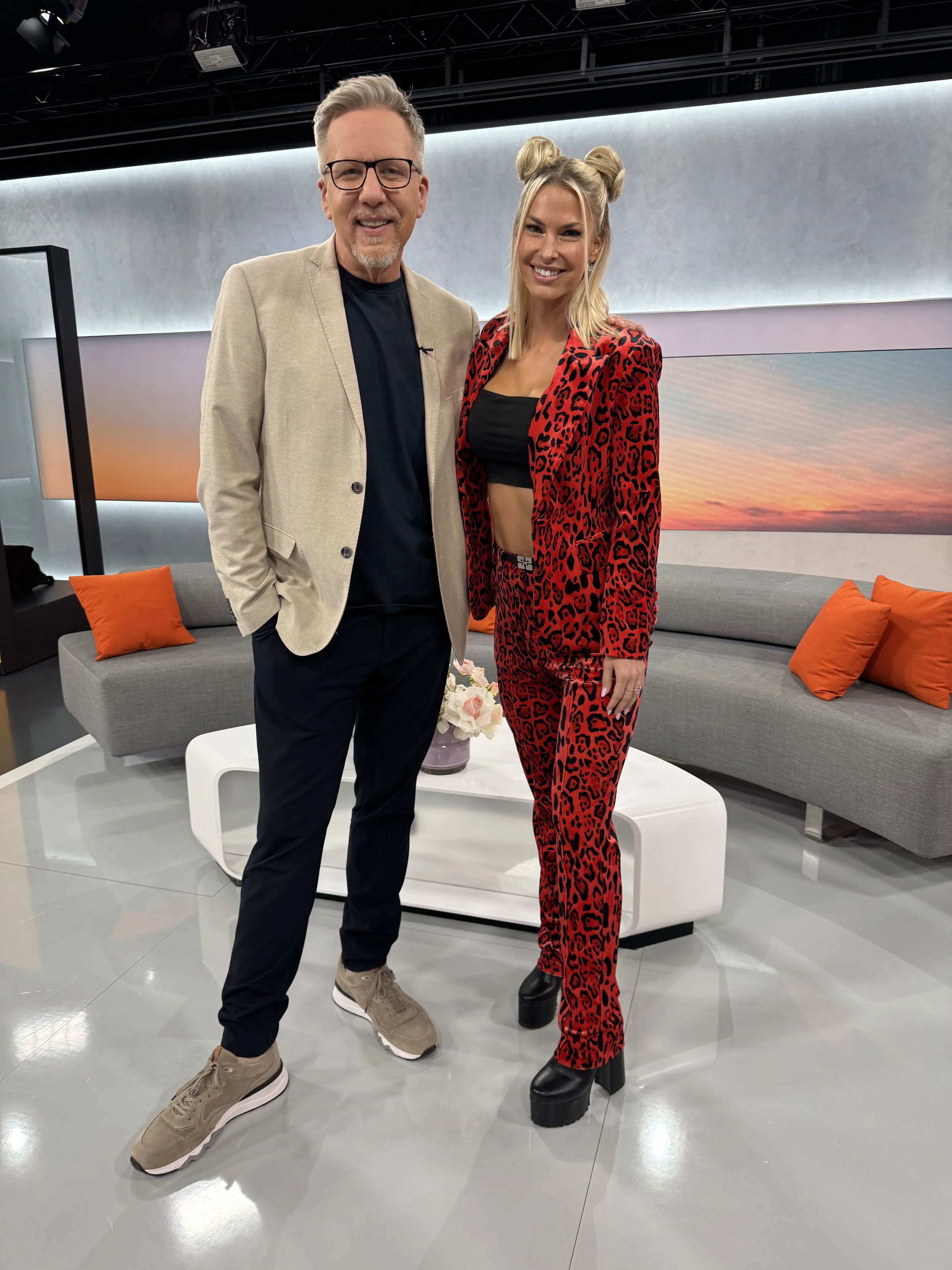 Zwei Menschen stehen in einer modernen Fernsehlounge, lächeln und posieren für die Kamera. Der Mann trägt eine beige Jacke, schwarze Hose und Turnschuhe, die Frau trägt ein rotes, leopardmusterndes Outfit mit Plateauschuhen.