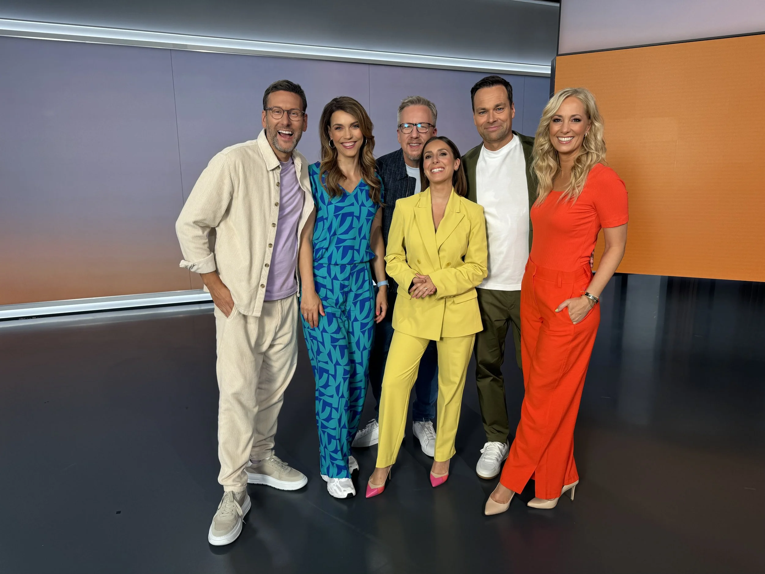 Sieben Menschen posieren zusammen in einer modernen TV-Studioset. Sie lachen und tragen bunte, modische Kleidung.