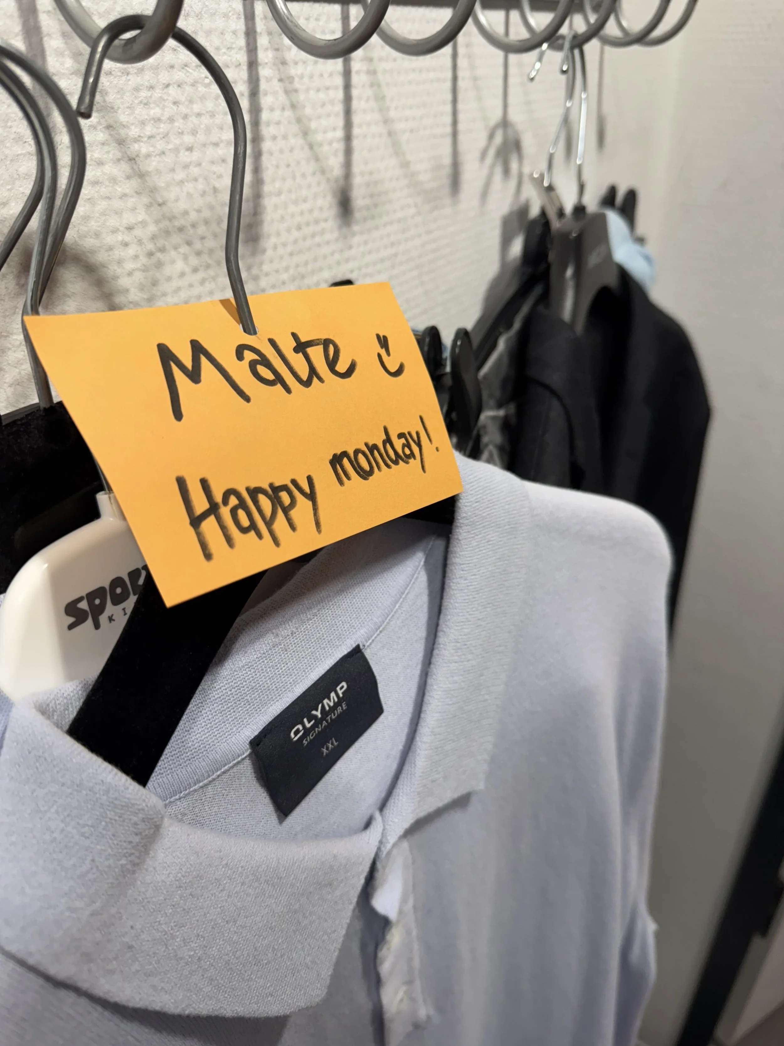 Herrenoberteil in Grau mit Marke Olymp auf einem Kleiderhaken, daneben ein orangefarbiger Zettel mit der Aufschrift »Malte :) Happy Monday!"