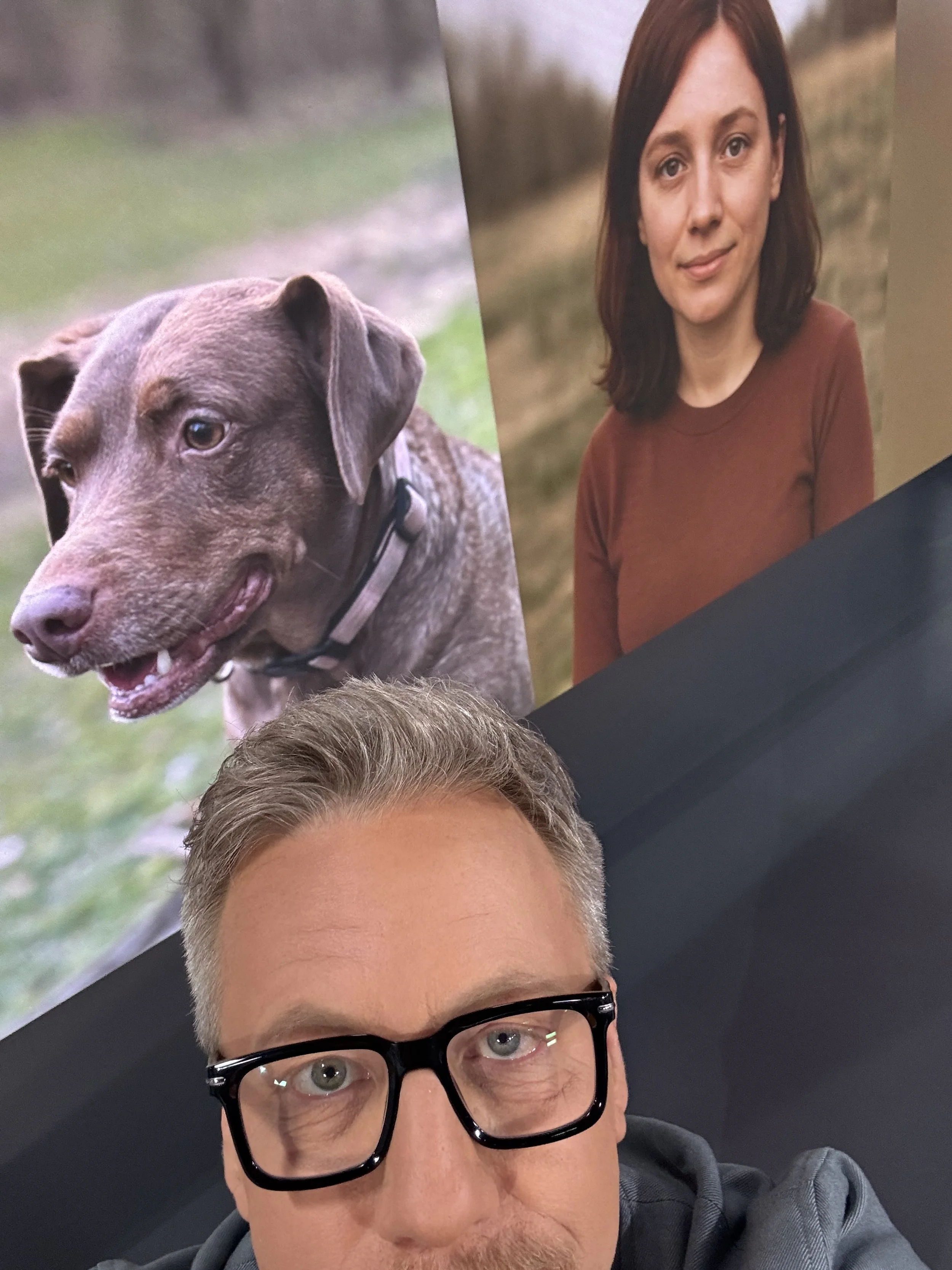 Nahaufnahme eines Mannes mit Brille, im Hintergrund ein Poster mit einem Hund und einer Frau, die auf dem Wandposter zu sehen sind.