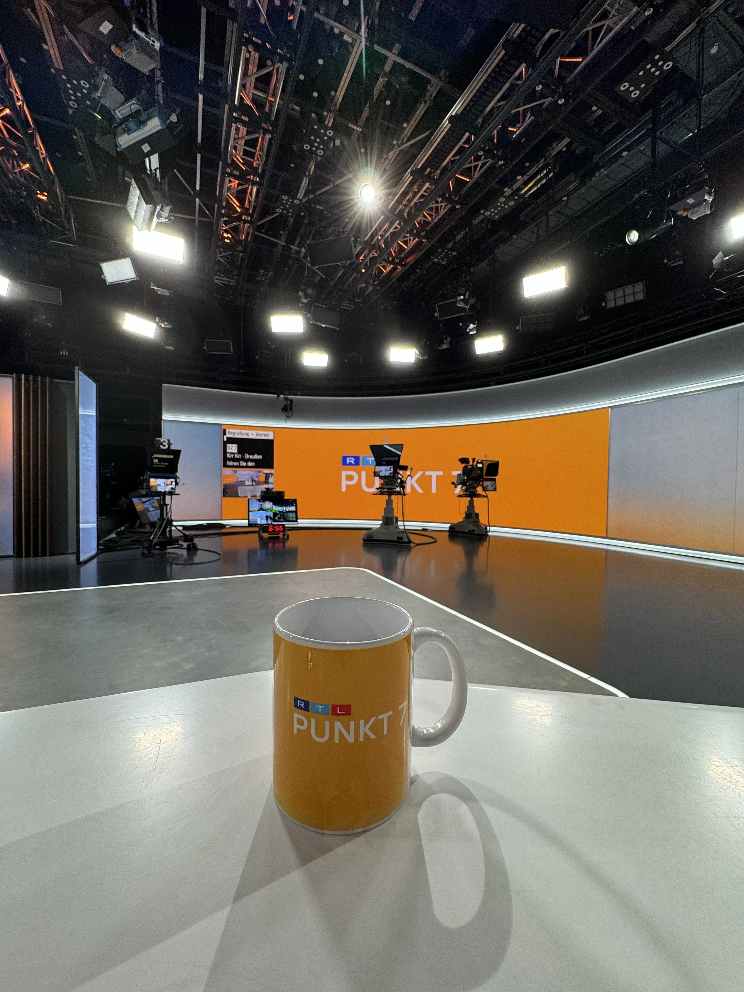 Studio-Set mit drei Kameras, beleuchtete Kulisse mit orange Hintergrund und den Worten 'RTL PUNKT 12' auf einem großen Bildschirm. Im Vordergrund eine Tasse mit dem RTL PUNKT 12 Logo auf einem weißen Tisch.