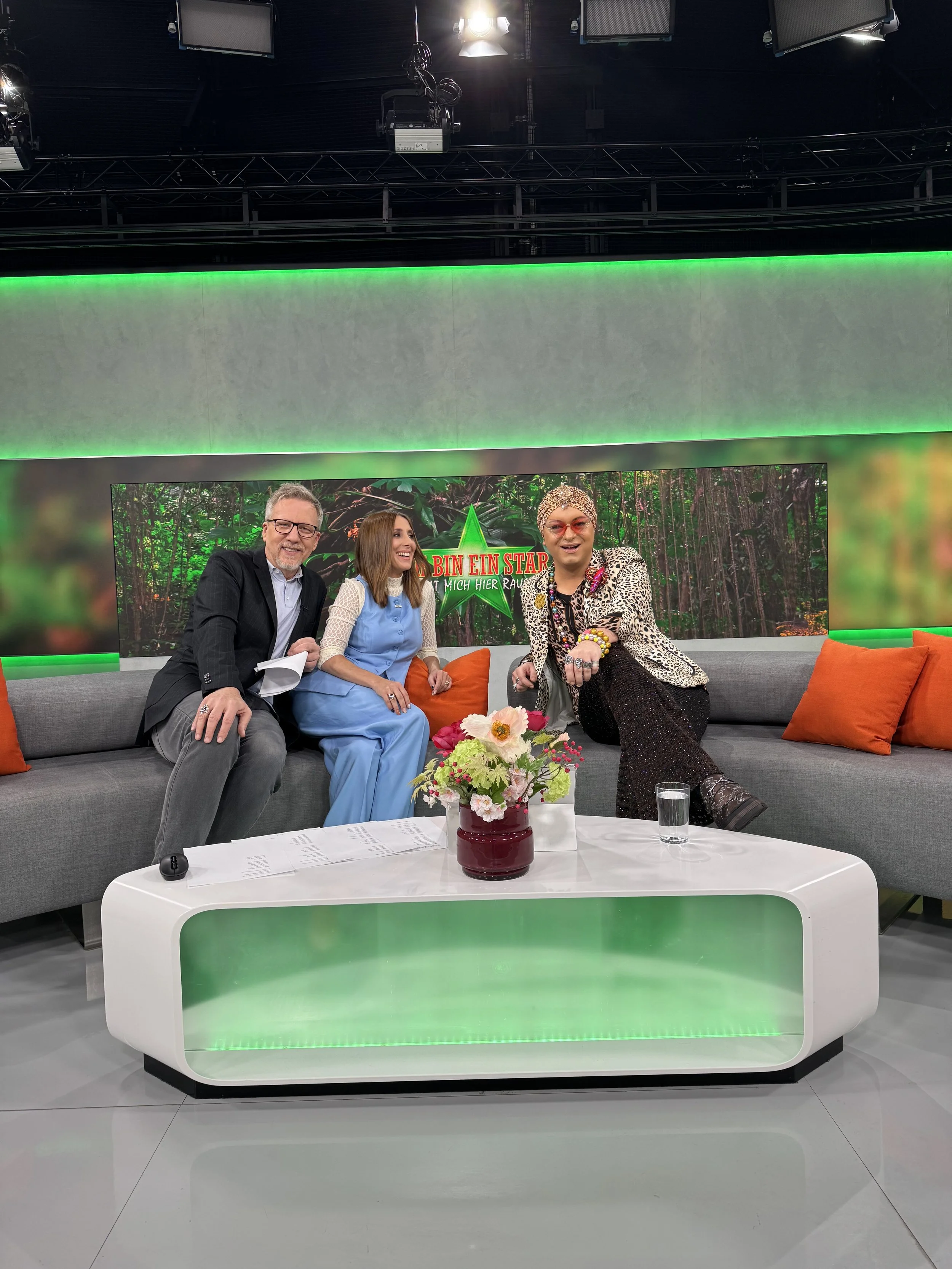 Drei Personen sitzen auf einer Couch in einem Fernsehstudio. Hinter ihnen ein Bildschirm mit einem Waldbild und einem grünen Stern mit roter Schrift. Auf dem Couchtisch liegt eine Blumenvasen mit Blumen, ein Glas Wasser und einige Papiere.