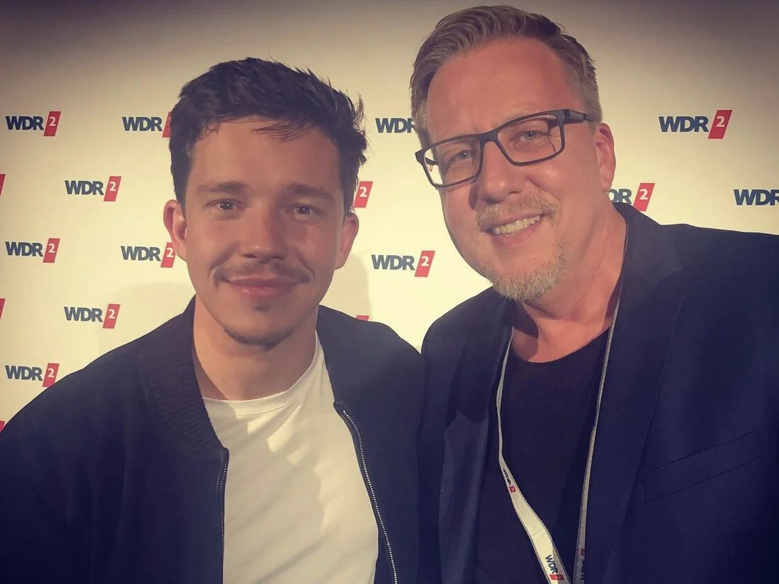 Zwei Männer posieren für ein Foto vor einem Hintergrund mit dem Logo von WDR 2.