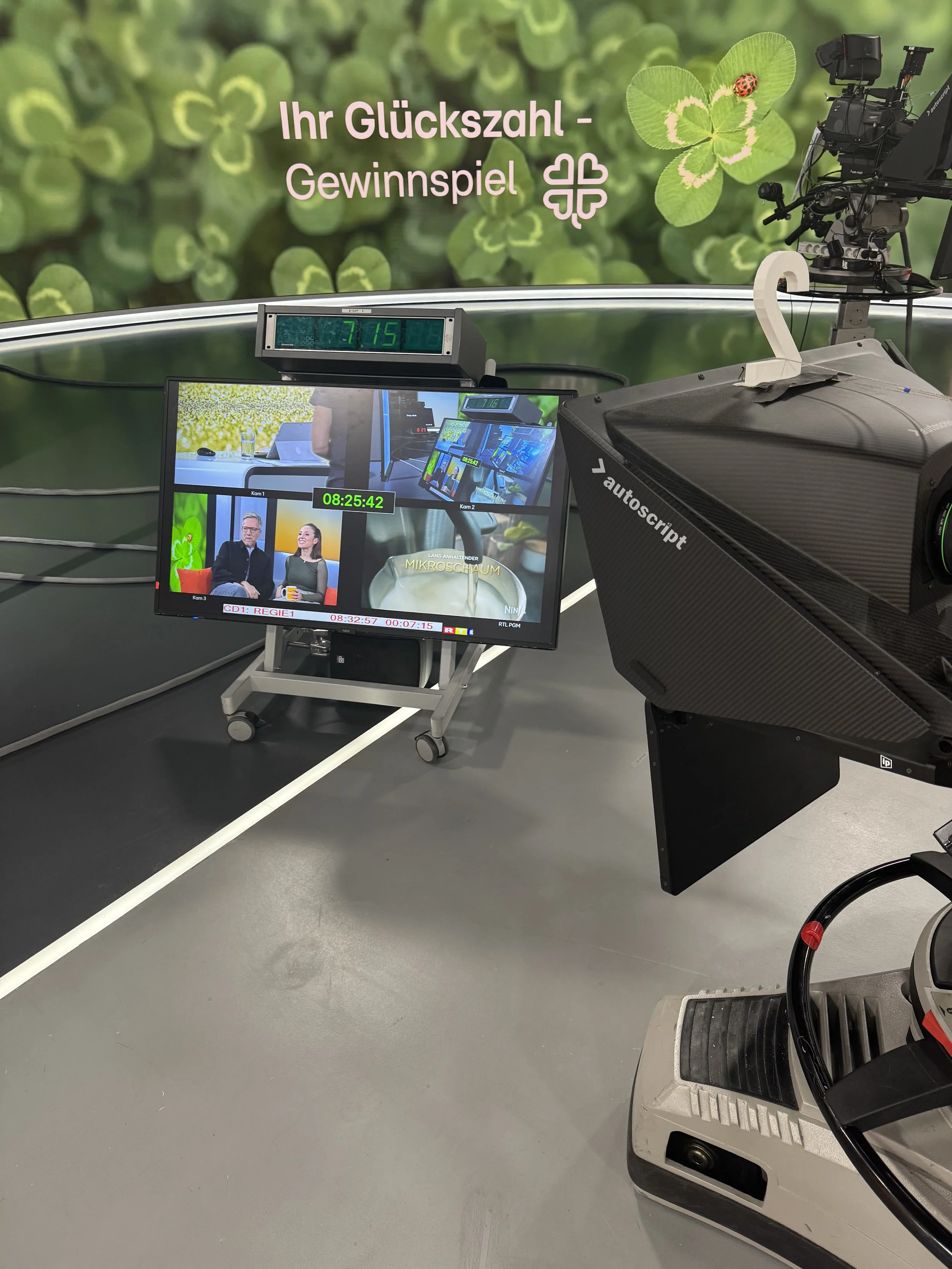 Aufnahme von einer Fernsehstudio-Produktionsumgebung mit Monitoren, Kameras und einem Hintergrund mit vierblättrigem Kleeblatt und Glückszahl, sowie einem Gewinnspiel-Logo.