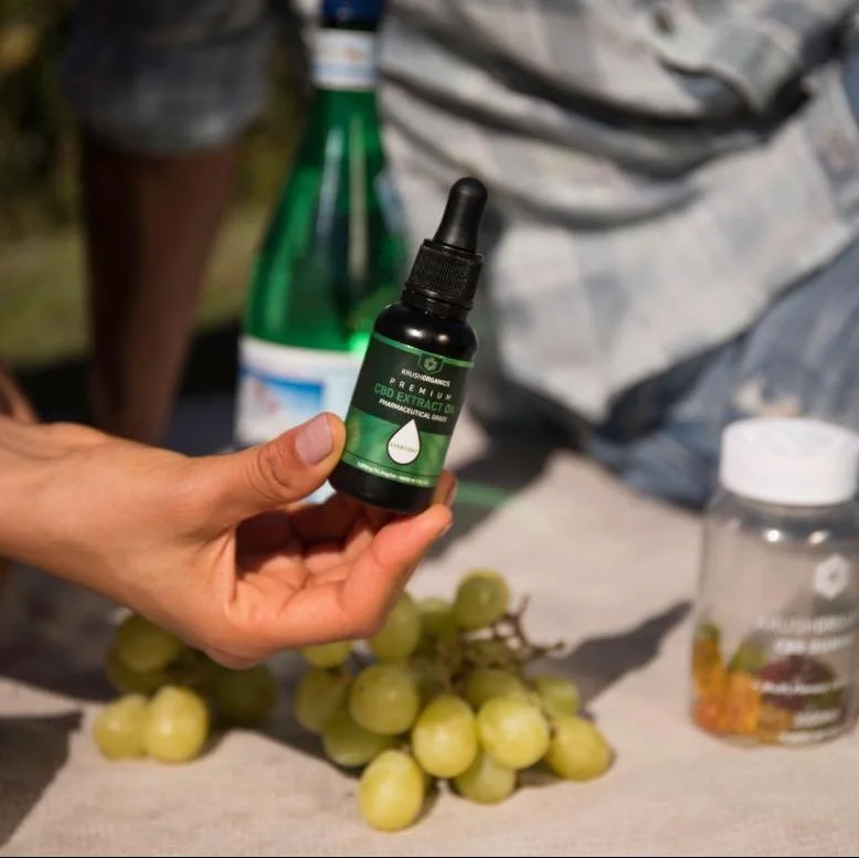 A person's hand holding a small black bottle labeled 'CBD Extract' with a dropper cap, in front of a bunch of green grapes and other bottles on a beige cloth surface.