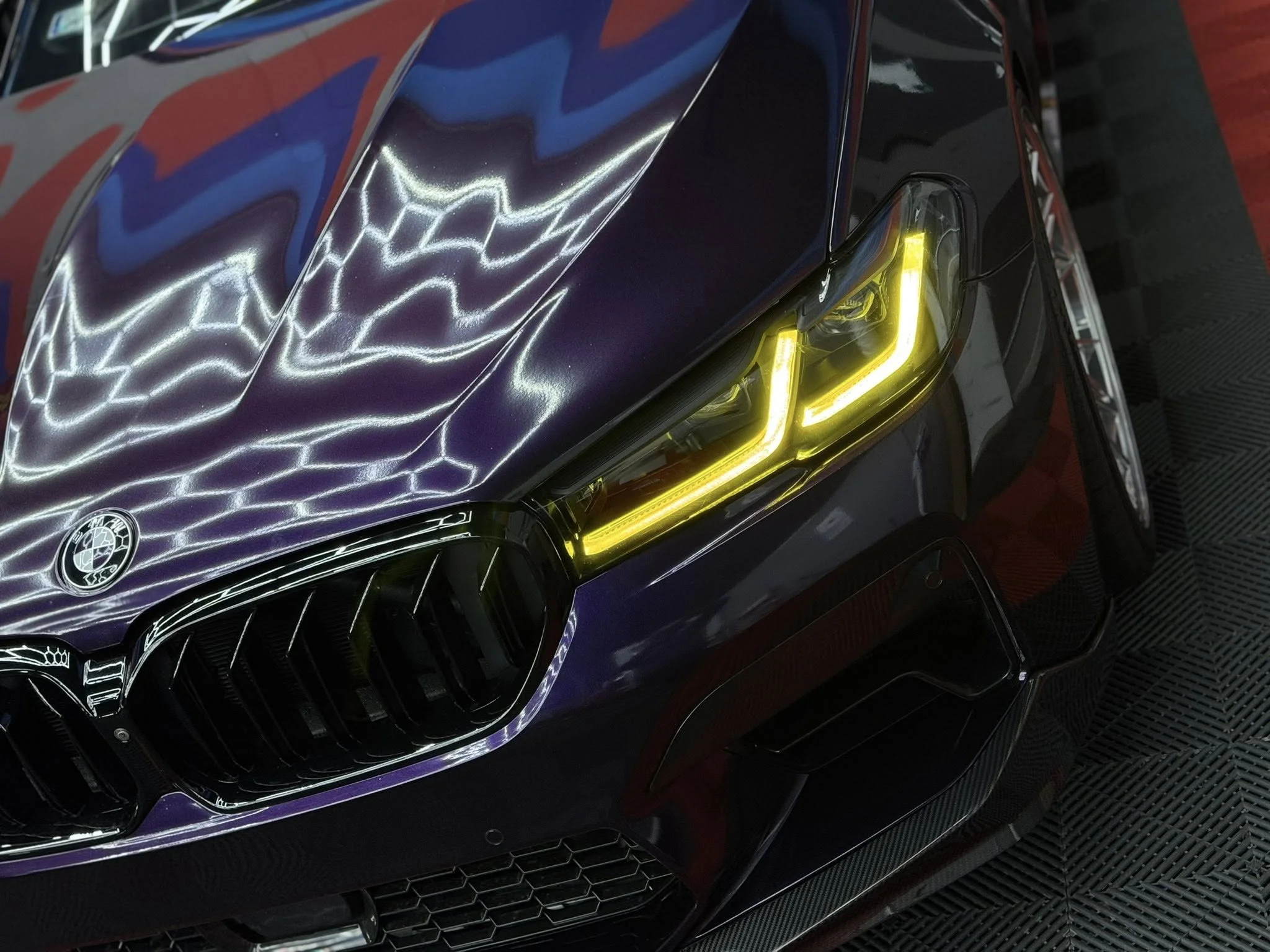 Close-up of a black luxury car's front, showcasing yellow LED headlights, a V8 badge, and a reflective hood with purple and blue hues.
