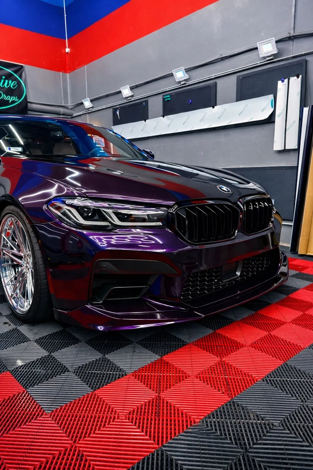A black BMW car with glossy finish parked in a garage with a red and black checkered floor. The garage wall has various tools and equipment mounted on it, and the ceiling features bright lighting.